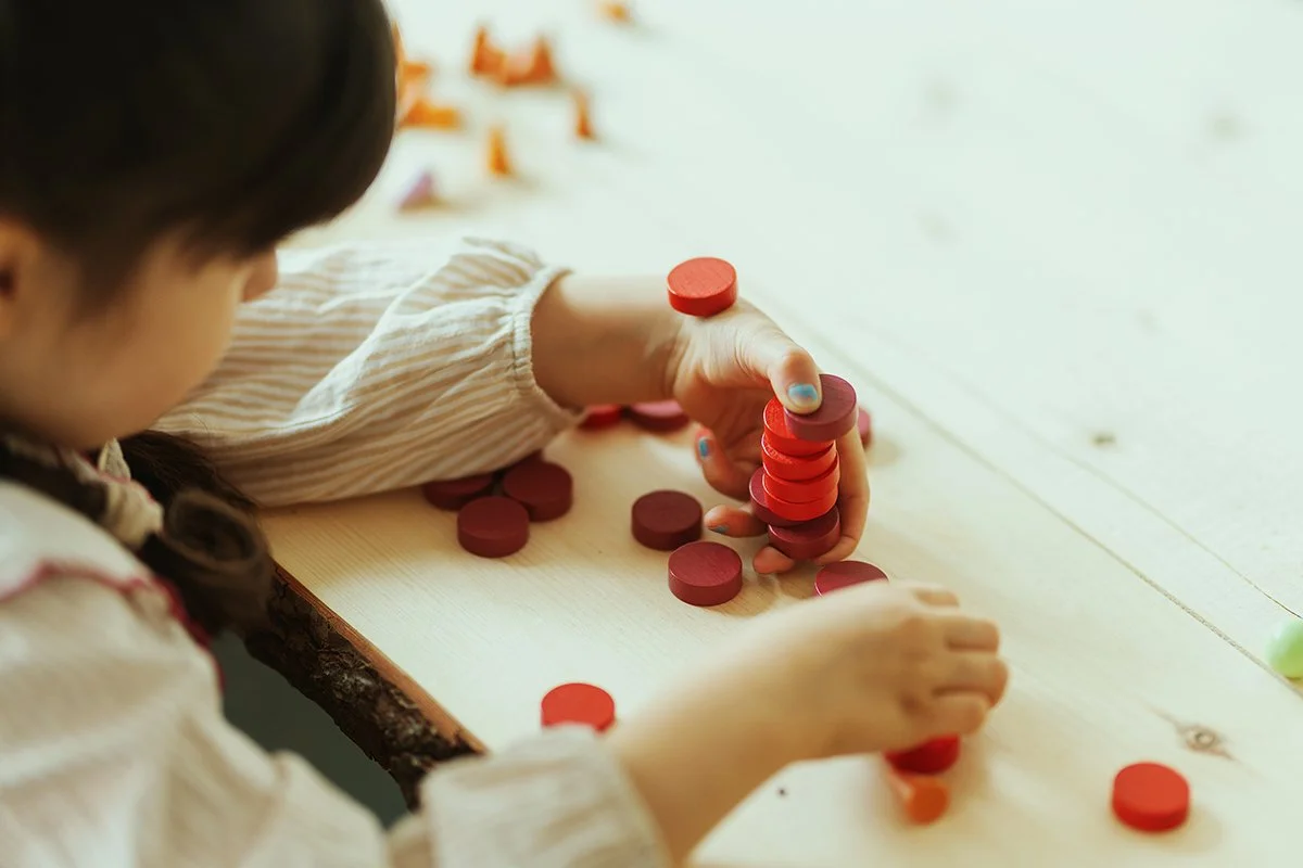 A young girl is playing with red wooden discs on a light-colored table, stacking them in a tower.