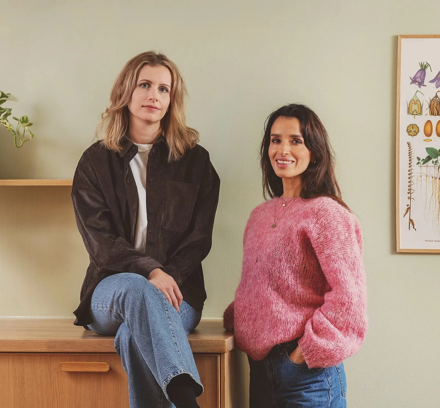 Two women with brown hair, one with wavy hair and the other with straight hair, pose indoors, one sitting on a wooden ledge and the other standing beside her, both smiling at the camera. The woman on the left wears a black jacket and blue jeans, while the woman on the right wears a pink sweater and dark jeans, with light green walls and framed botanical art in the background.