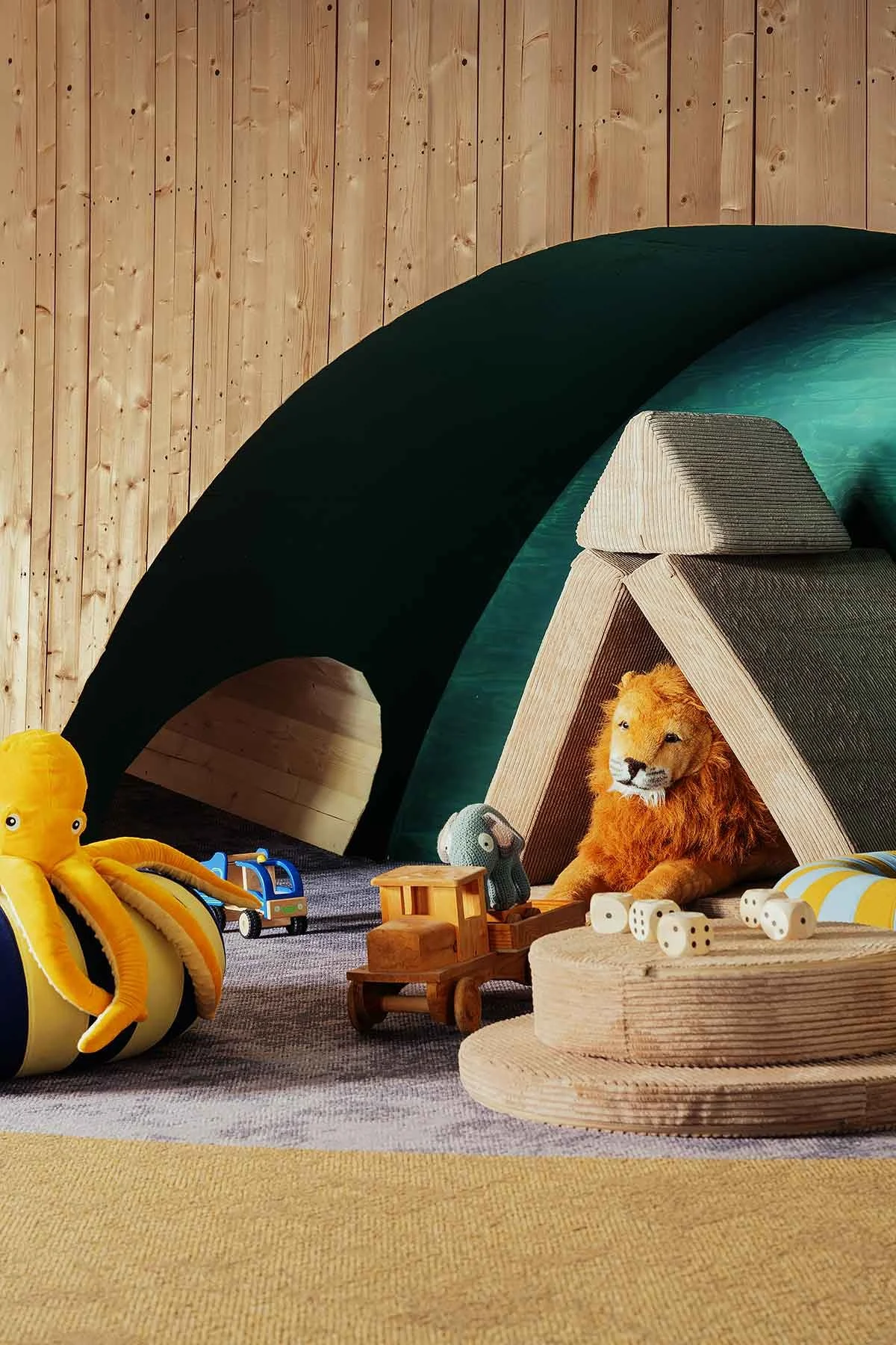 Children's playroom with a wooden wall, a black foam tunnel, a wooden teepee tent with a stuffed lion inside, and various toys including plush octopus, toy truck, crocheted octopus, dice, and wooden blocks.