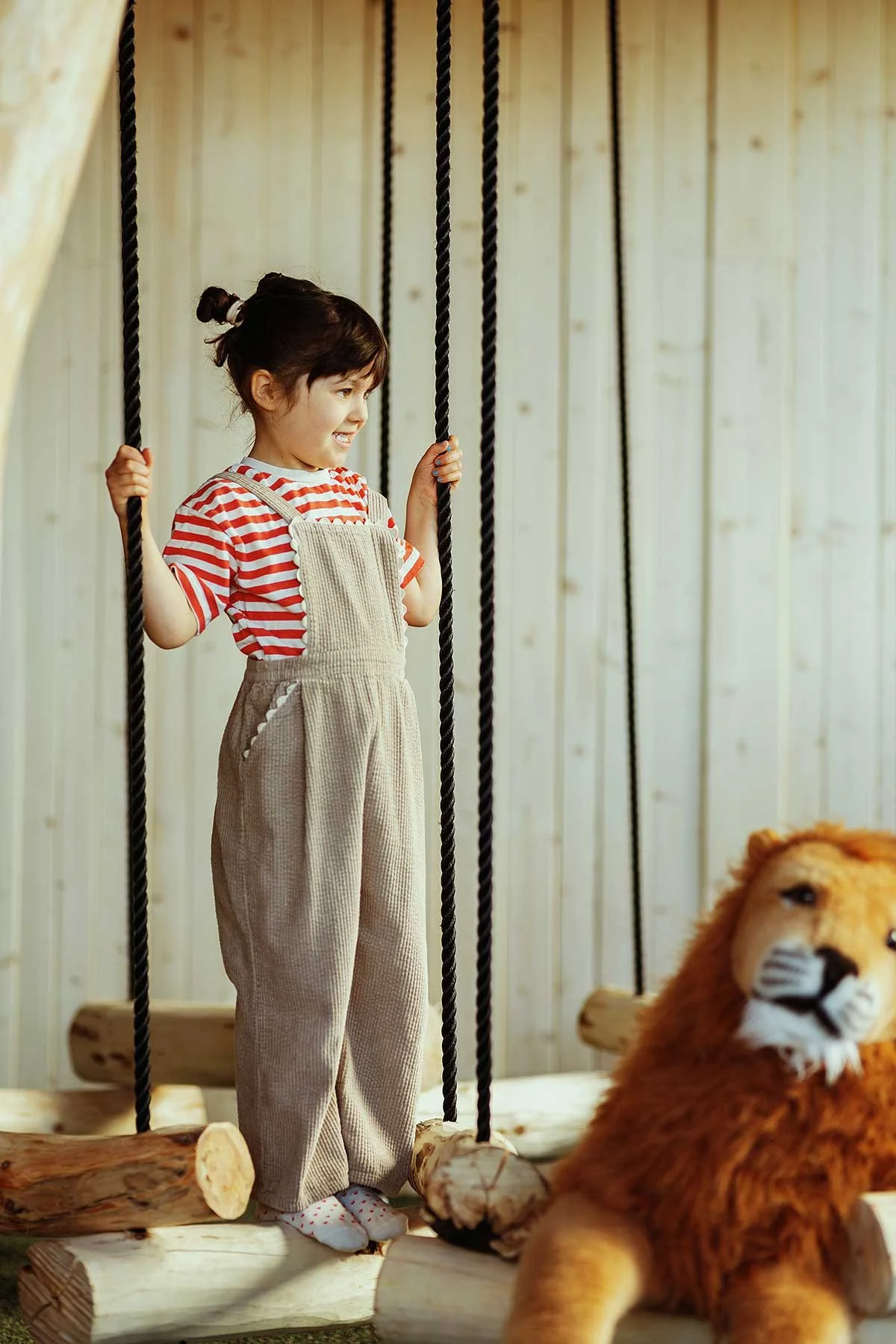 A young girl with dark hair in a bun, wearing a red and white striped shirt, beige overalls, and socks with red dots, standing on a wooden swing set, looking at a plush lion toy in a cozy indoor space with wooden wall paneling.