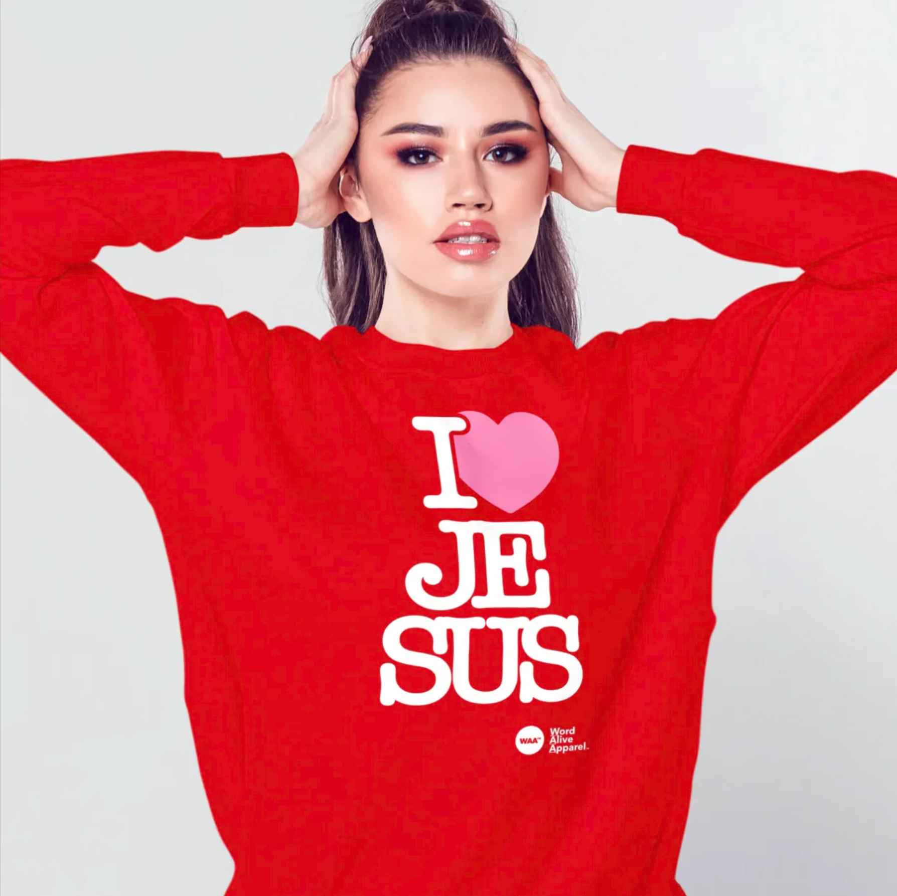 A woman with dark hair wearing a red sweatshirt with the text 'I ❤️ JEE SUS' in white and pink letters. She has her hands on her head, and the background is plain and light-colored.