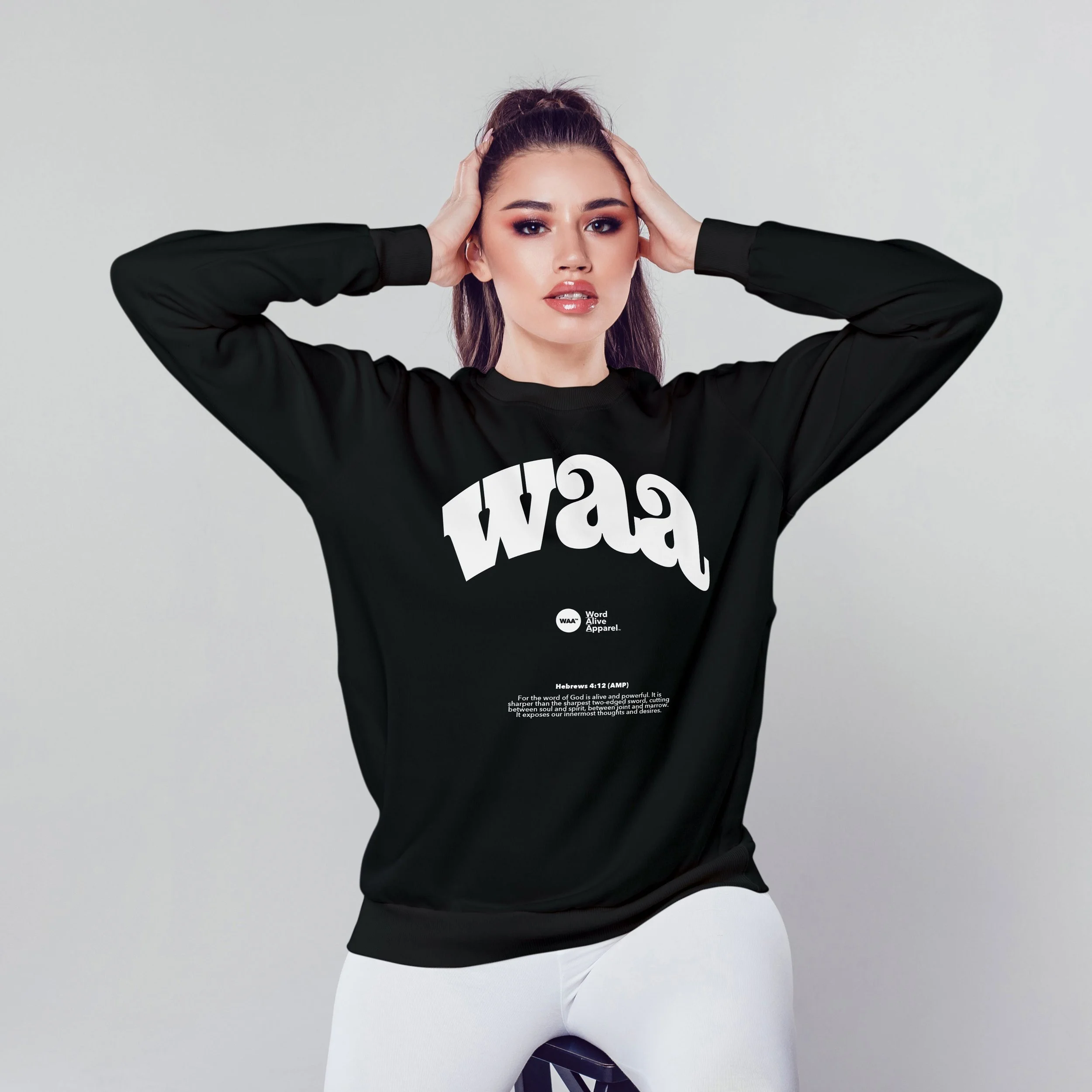 Young woman with dark hair and makeup, wearing a black sweatshirt with a white printed design, posing with her hands on her head against a light gray background.