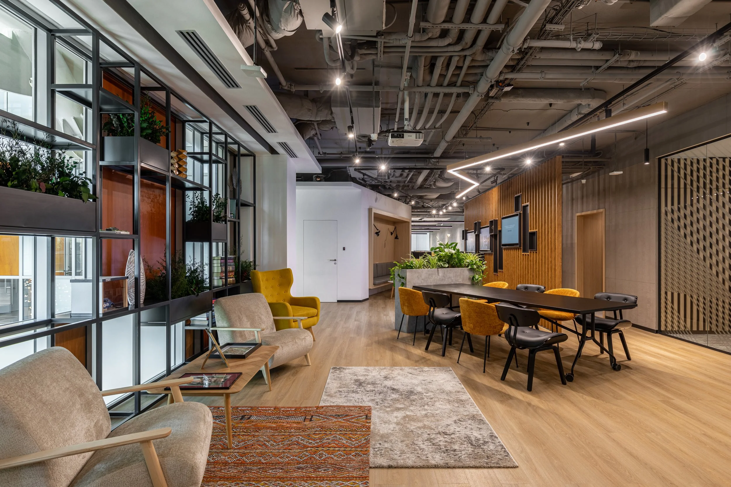 Modern office lounge with armchairs, tables, a long dining table with chairs, potted plants, wall TVs, and a decorative ceiling, illuminated by track and strip lighting.