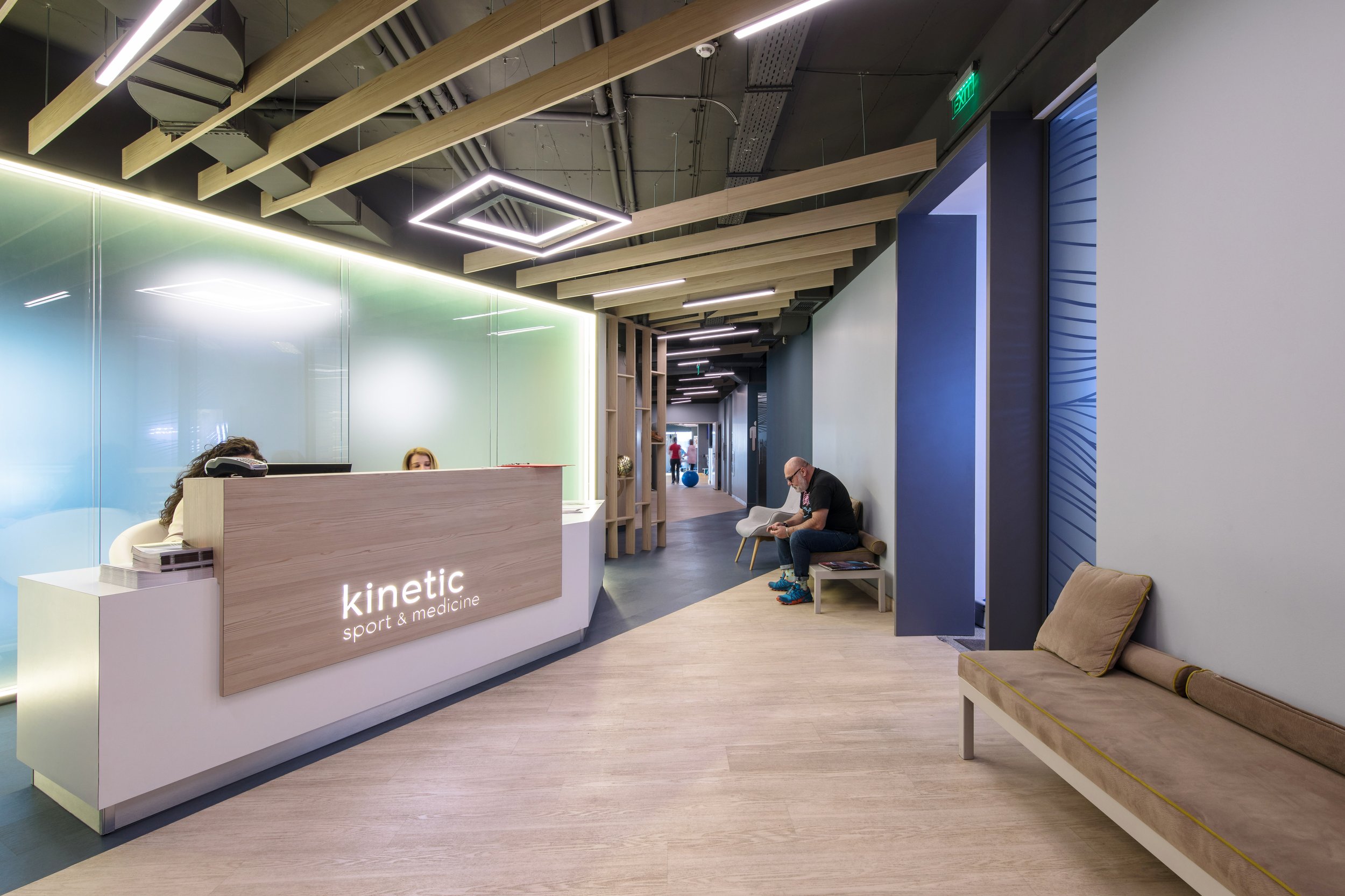 Modern medical clinic reception area with a wooden reception desk, a man seated on a bench reading, and a few people walking in the hallway.