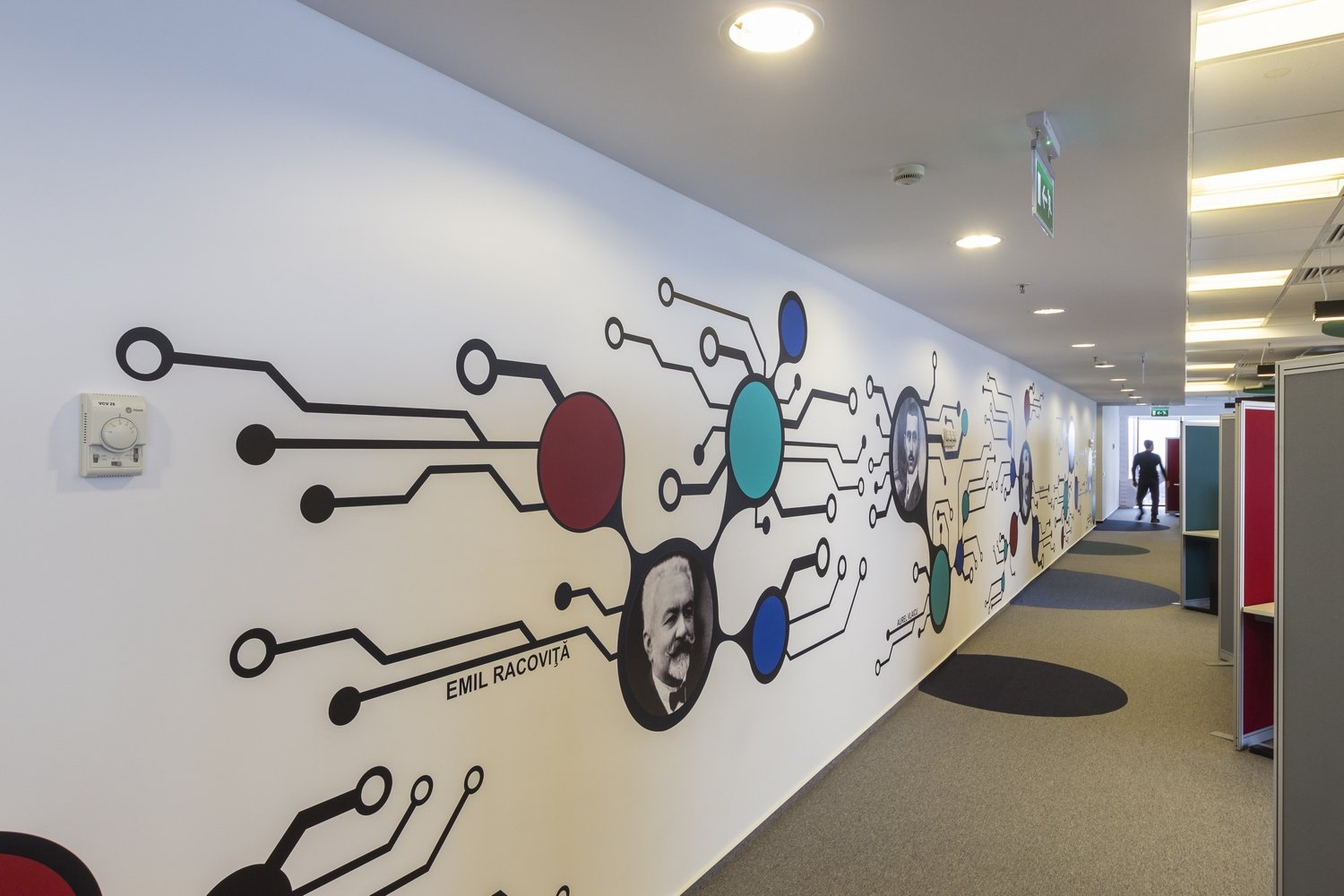 A hallway with a white wall featuring a large, colorful mosaic-style mural of interconnected circles and lines with a black and white portrait of Emil Racoviță in the center. The ceiling has recessed lighting, and there are some people at the far end of the hallway.