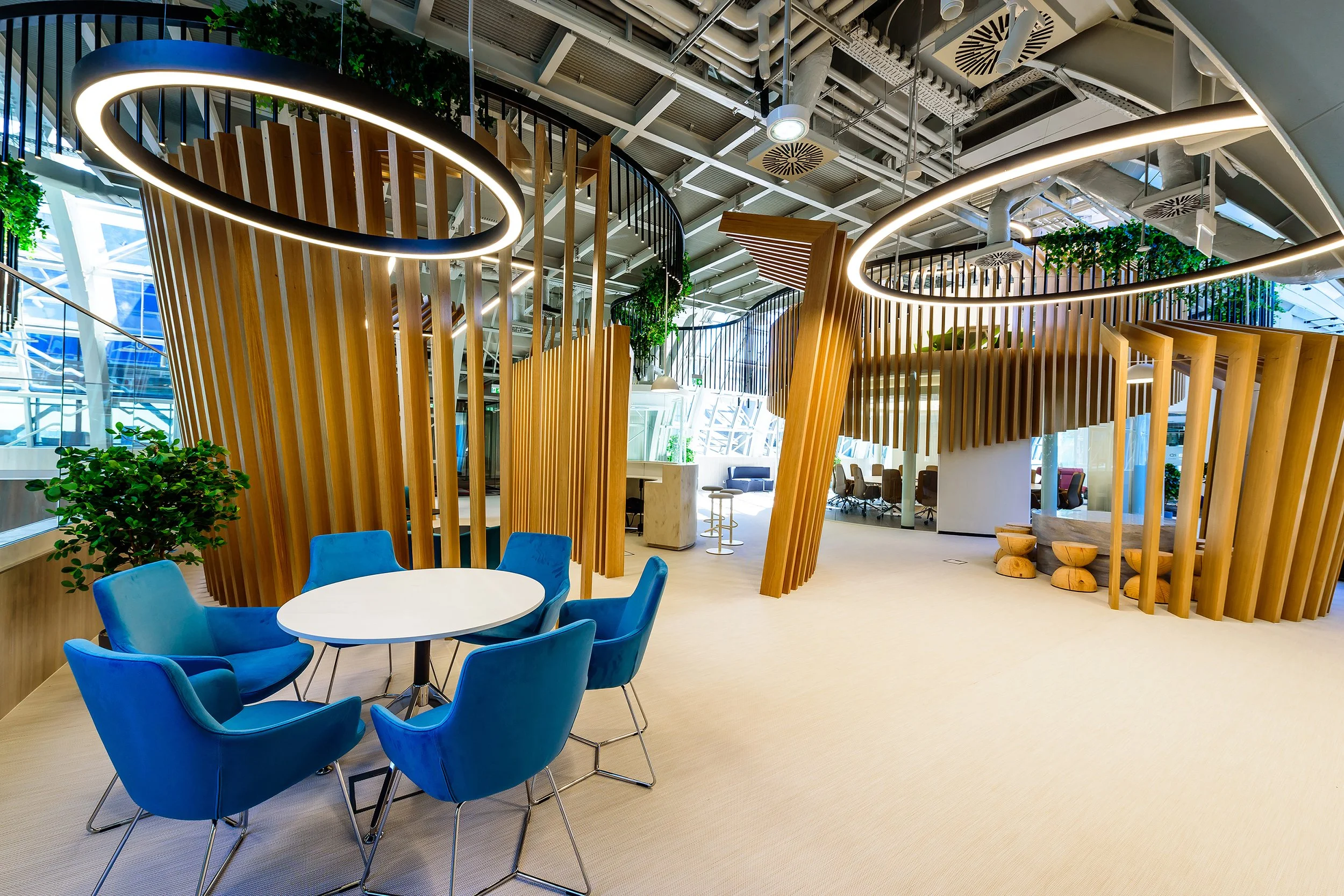 Modern office space with wooden structures, circular light fixtures, blue chairs around a white table, and greenery.