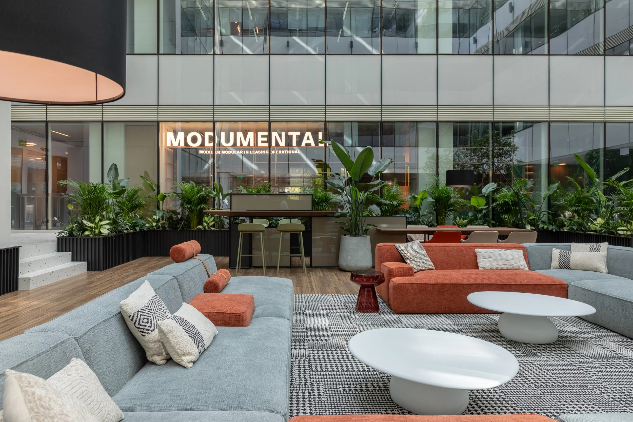 Modern indoor lounge area with gray and orange sofas, decorative pillows, round white coffee tables, and a large green potted plant, with a glass facade and a business sign in the background.