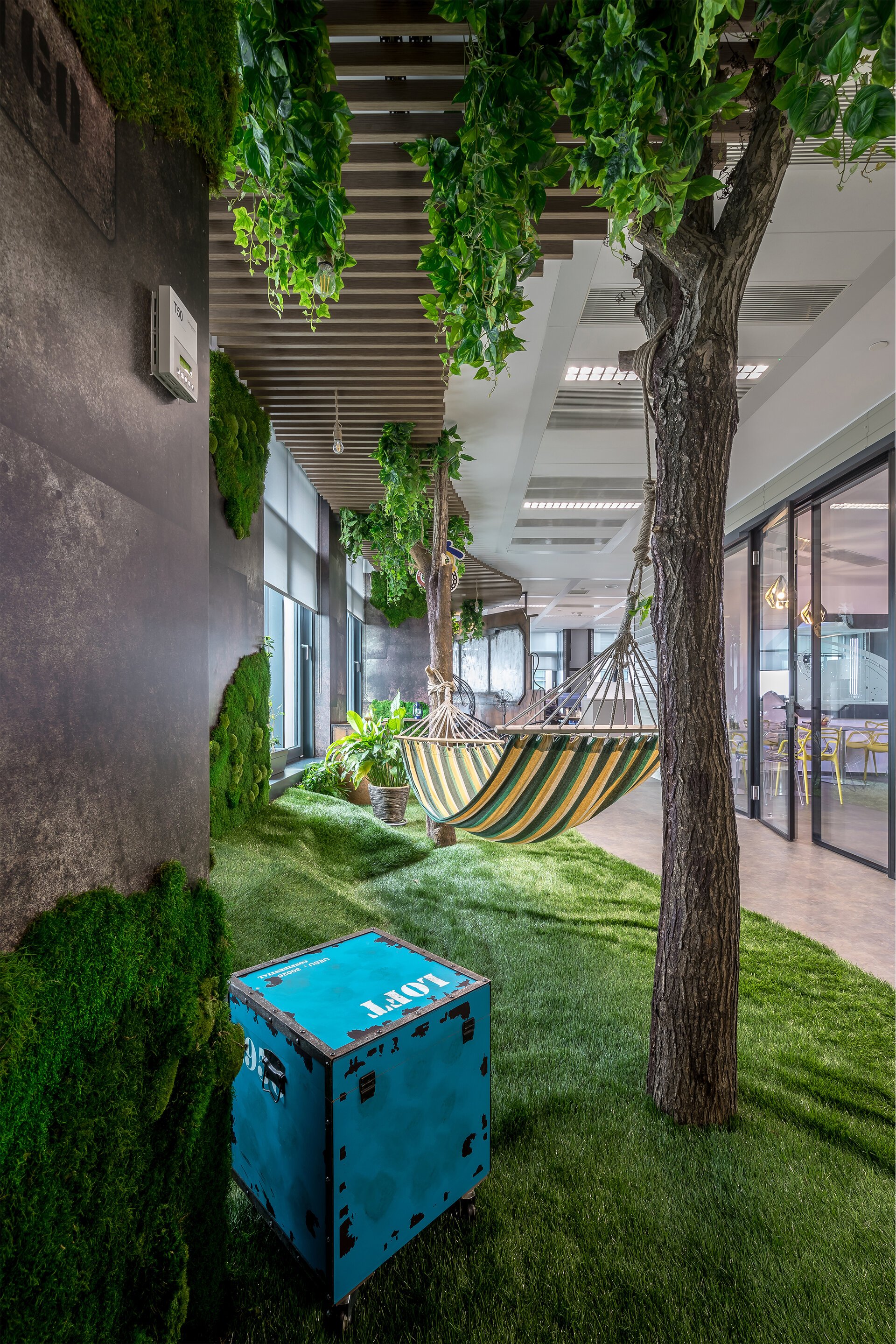 Indoor space with artificial grass, trees, hanging hammock, potted plants, and green wall accents, with windows and a sliding glass door.