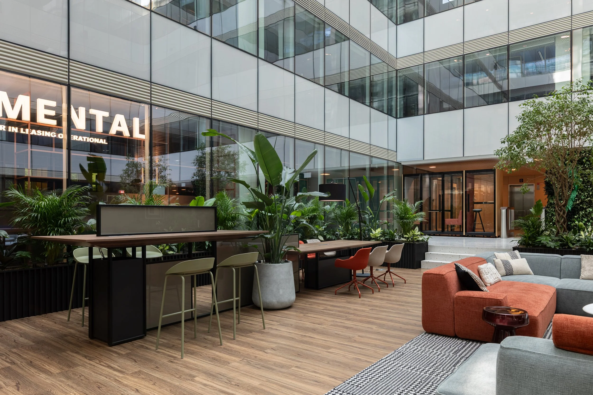 Indoor lounge area with wooden flooring, green plants, a gray and orange sectional sofa, a small round table, and high-top tables with chairs, with large glass windows and a modern office building facade in the background.