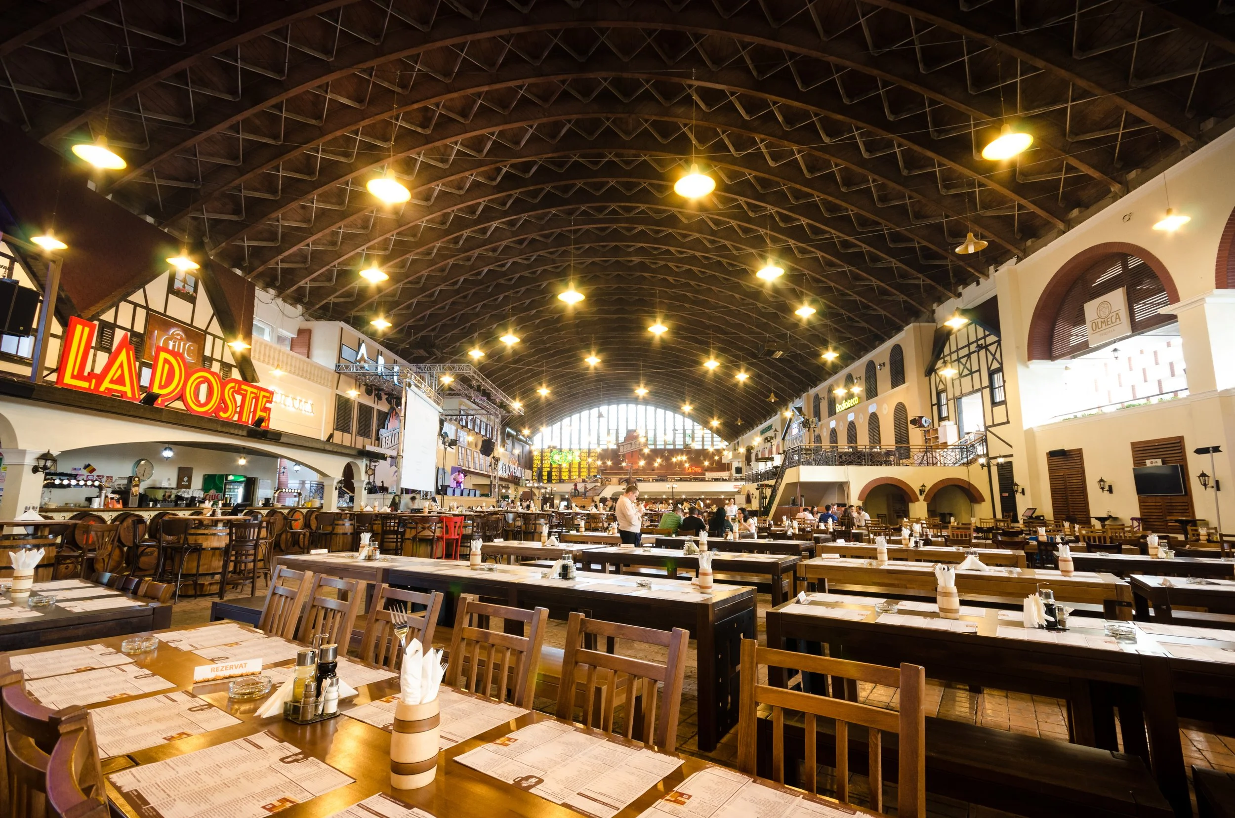 Spacious indoor restaurant with wooden tables and chairs, large arched windows, warm yellow lighting, and a high dark wooden ceiling, featuring signs like "La Poste" and various decorative elements.