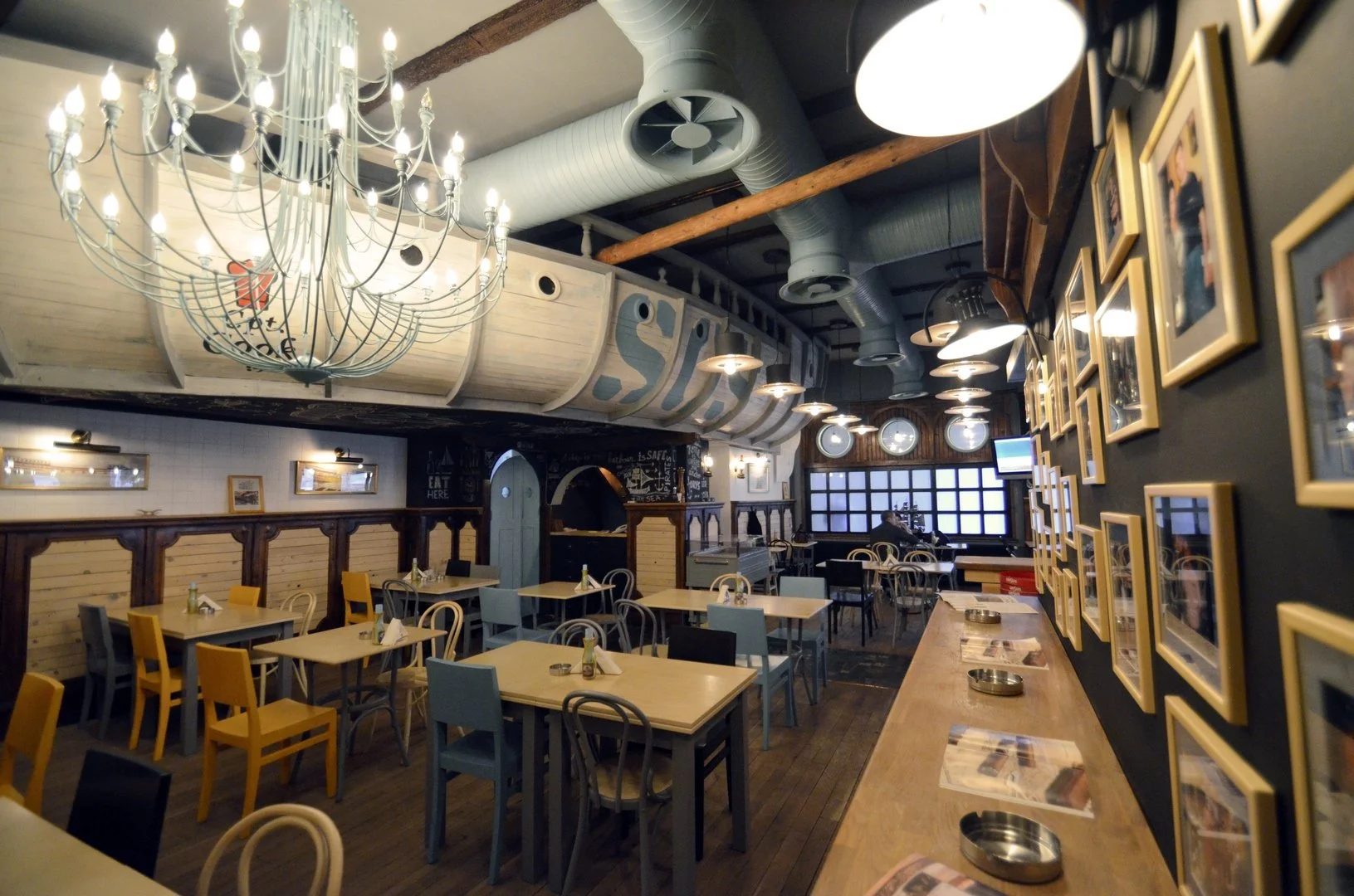 Interior of a restaurant with wooden tables, yellow and light blue chairs, framed photos on a dark wall, a large chandelier, and a bar area with placemats and metal ashtrays on the table.