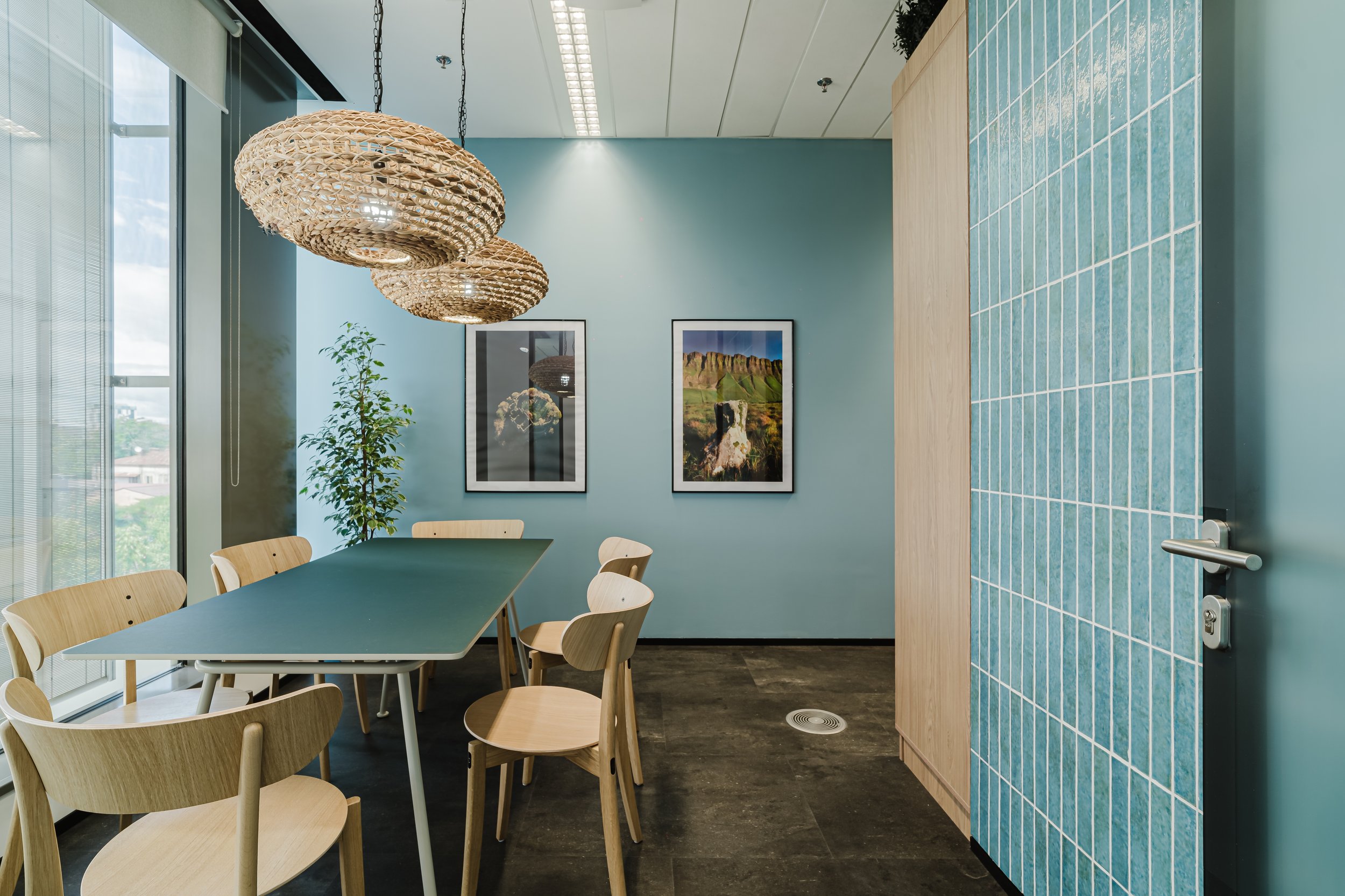 An interior conference room with a large window, light wood chairs around a rectangular table, wall art photos, woven pendant lights, a potted plant, and a partially visible blue tiled wall.