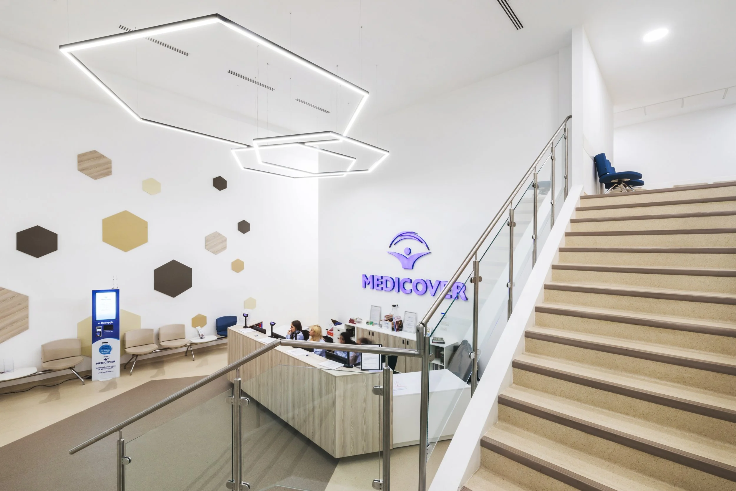 Reception area of a medical facility with a white counter, computers, and staff, illuminated ceiling lights, decorative wall with hexagonal panels, and a staircase leading to an upper level, featuring a "MEDICOVER" sign on the wall.
