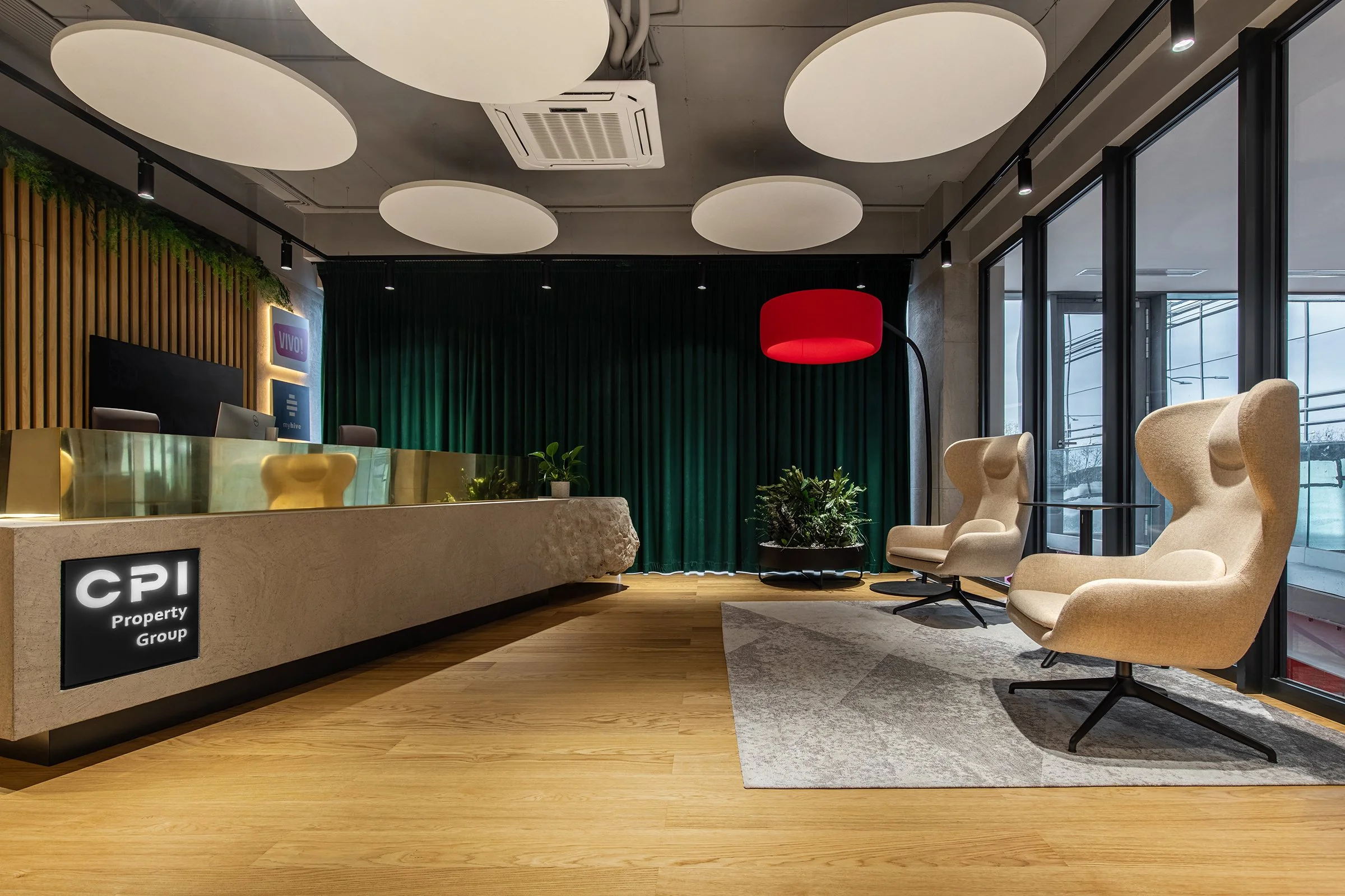 Modern office reception area with a beige counter, green curtain backdrop, armchairs, potted plants, red floor lamp, and large windows.