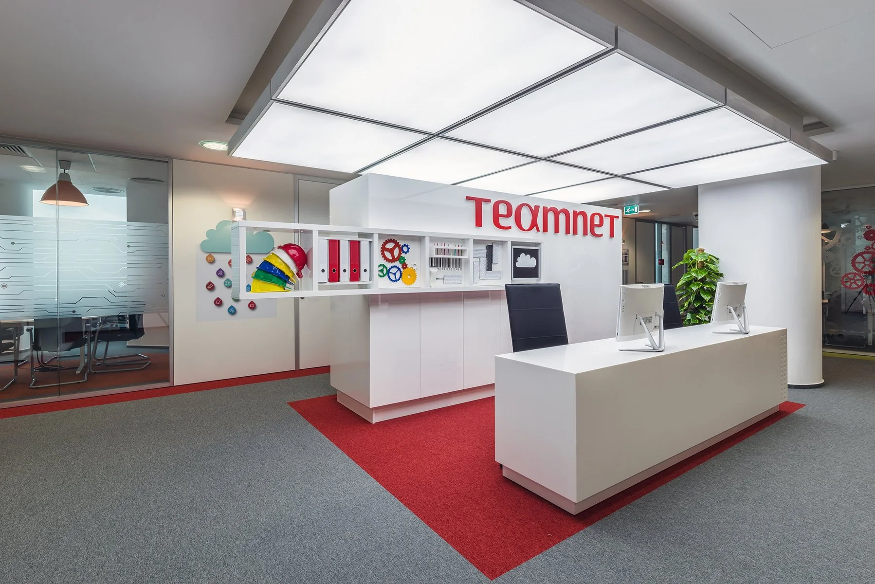 Modern office reception area with white desk, computer monitors, black chairs, and decorative wall art. The company's logo, 'TEAMNET,' is displayed on the wall.