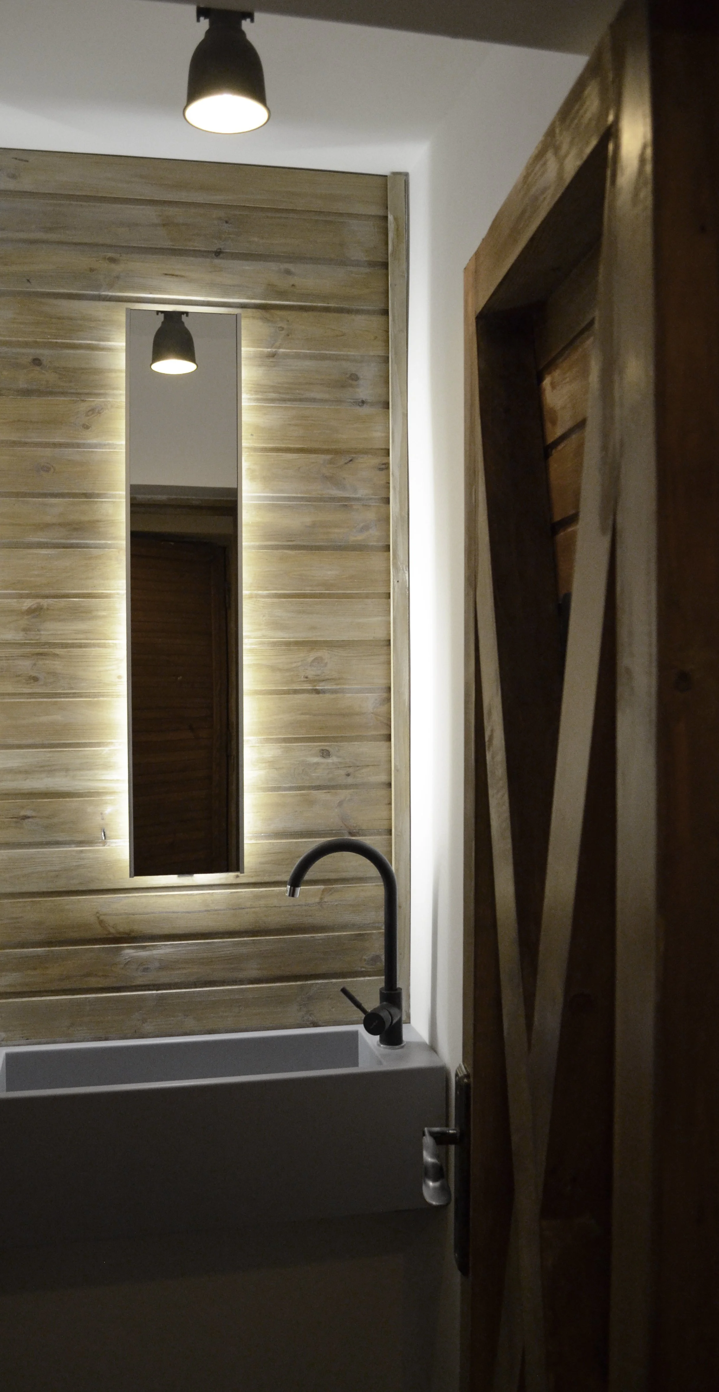 A bathroom interior with a black ceiling light, a mirror with backlighting, a wooden wall, a white sink with a black faucet, and a wooden door.