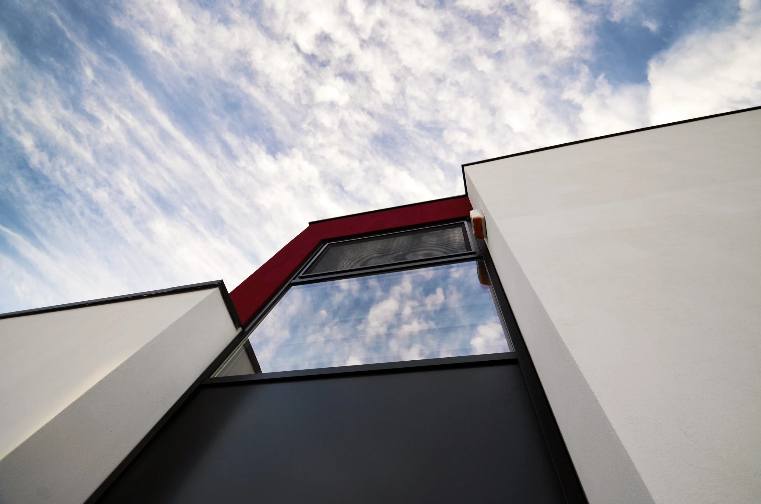 Low-angle view of a modern building with white and red exterior walls, a large window reflecting the sky with clouds, against a blue sky with wispy clouds.
