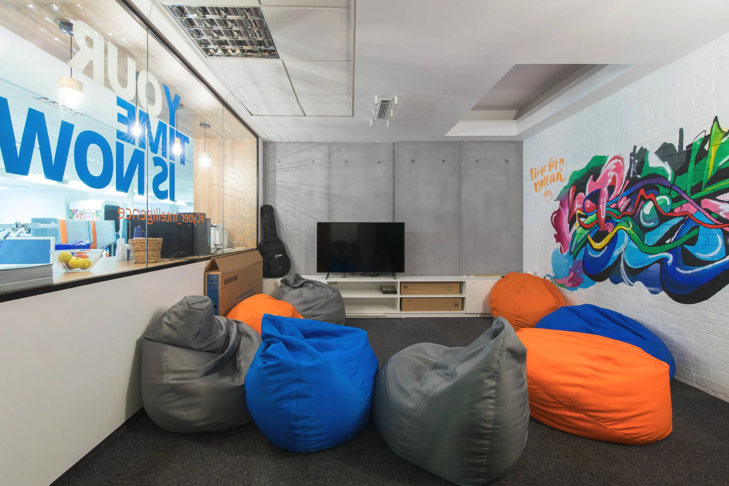 Indoor lounge area with colorful bean bags including orange, blue, and gray, a small flat-screen TV on a white entertainment unit, a musical instrument case, and a large vibrant mural on the white brick wall with colorful abstract designs and the words "Tune for a Break." There is a large window with text "YOUR TIME IS NOW" and miscellaneous items like a bowl of fruit and a cardboard box near the window.