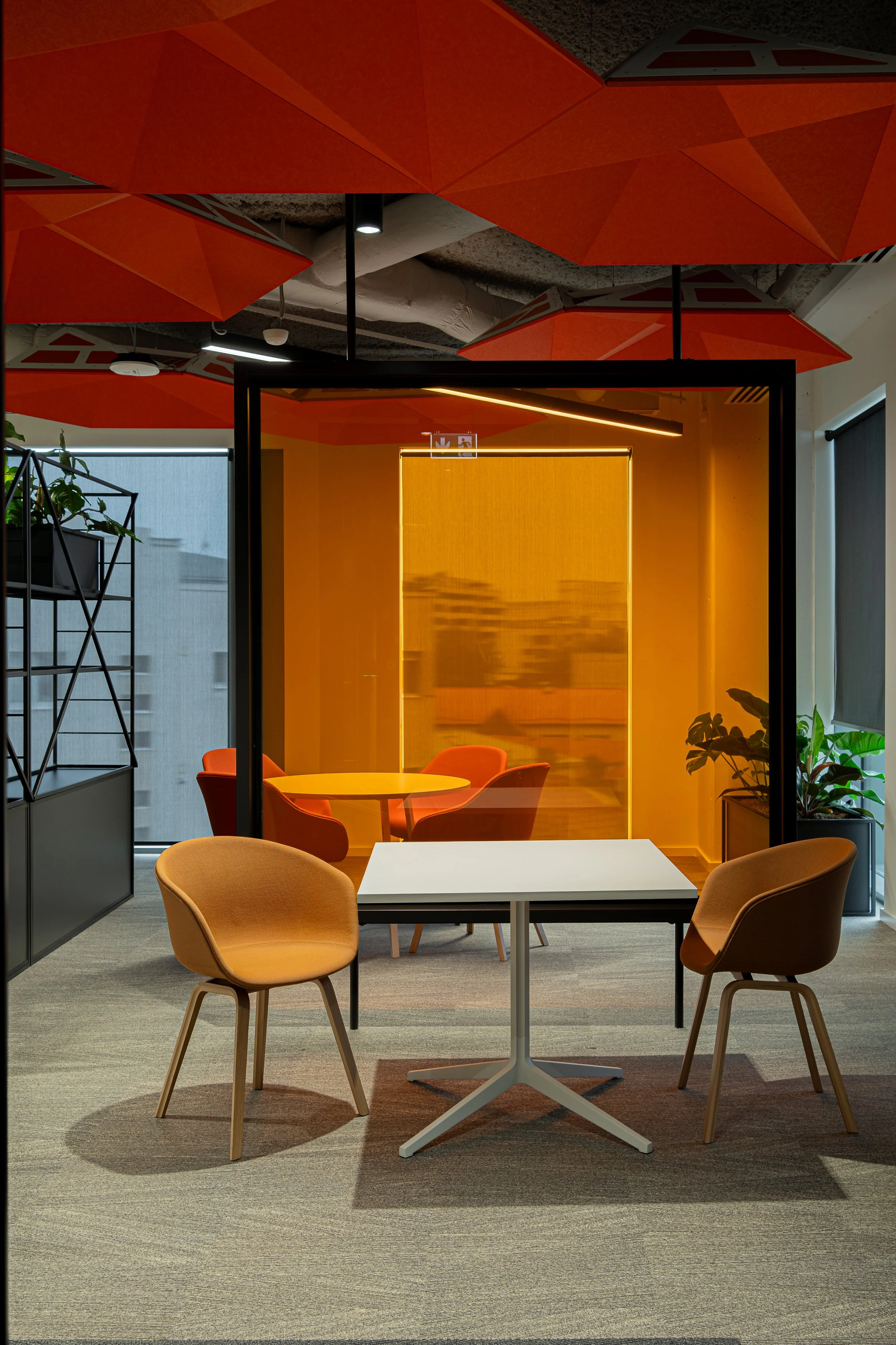 Modern office meeting area with two beige chairs, a white table, and a glass-enclosed room with orange walls and chairs inside. Large windows with city view and indoor plants.