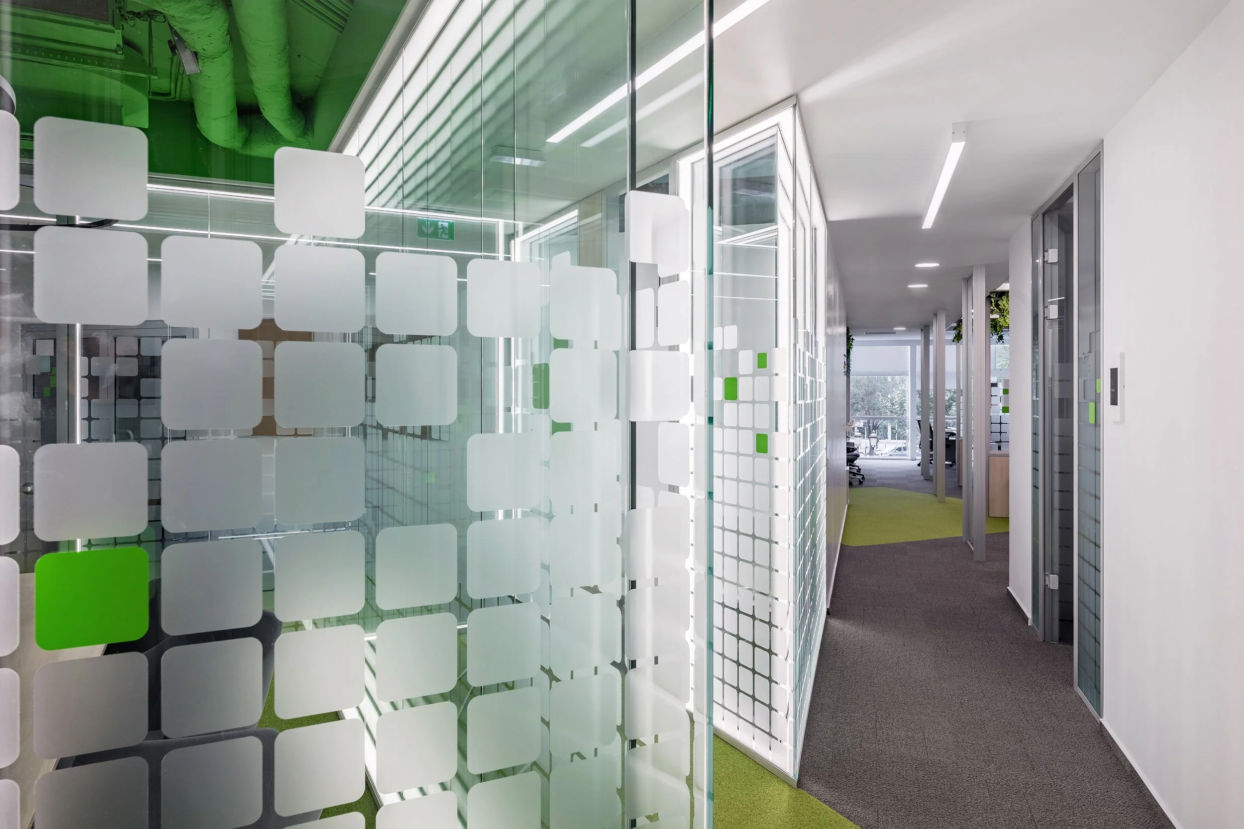 Modern office hallway with glass partitions and white walls; some partitions have frosted square patterns, and there is a mix of green and gray carpeted flooring.
