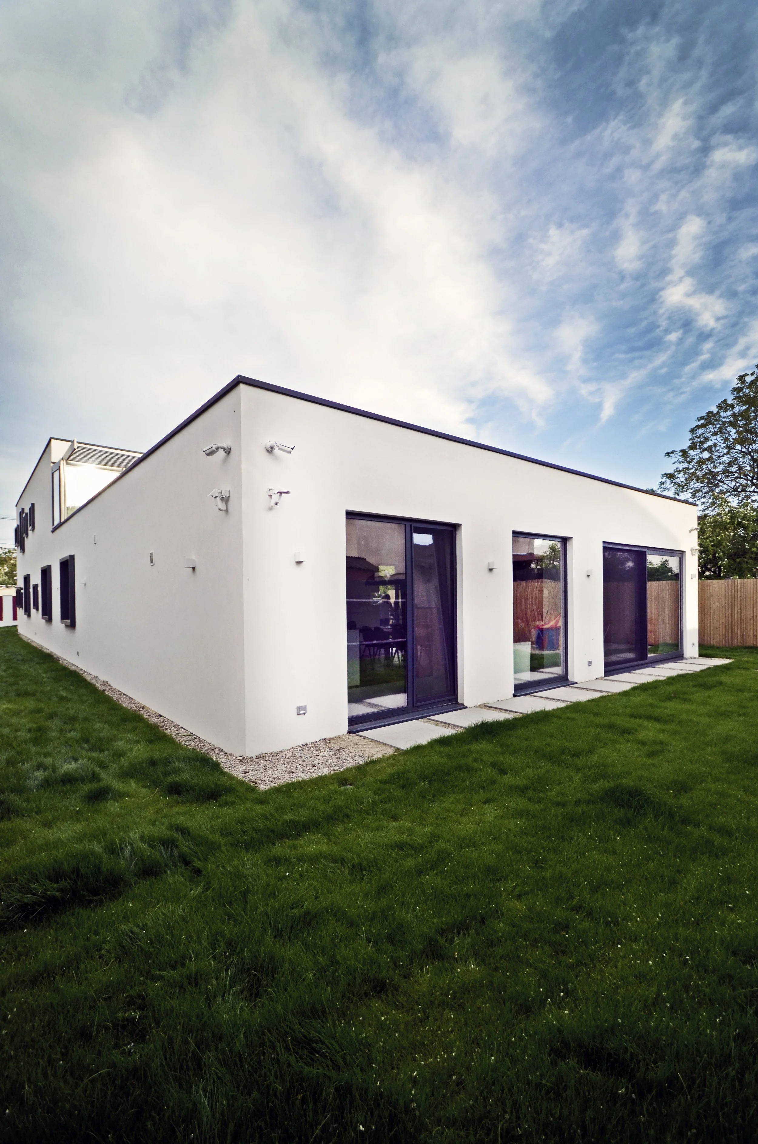 Modern white house with large sliding glass doors, a flat roof, and a well-maintained green lawn under a partly cloudy sky.