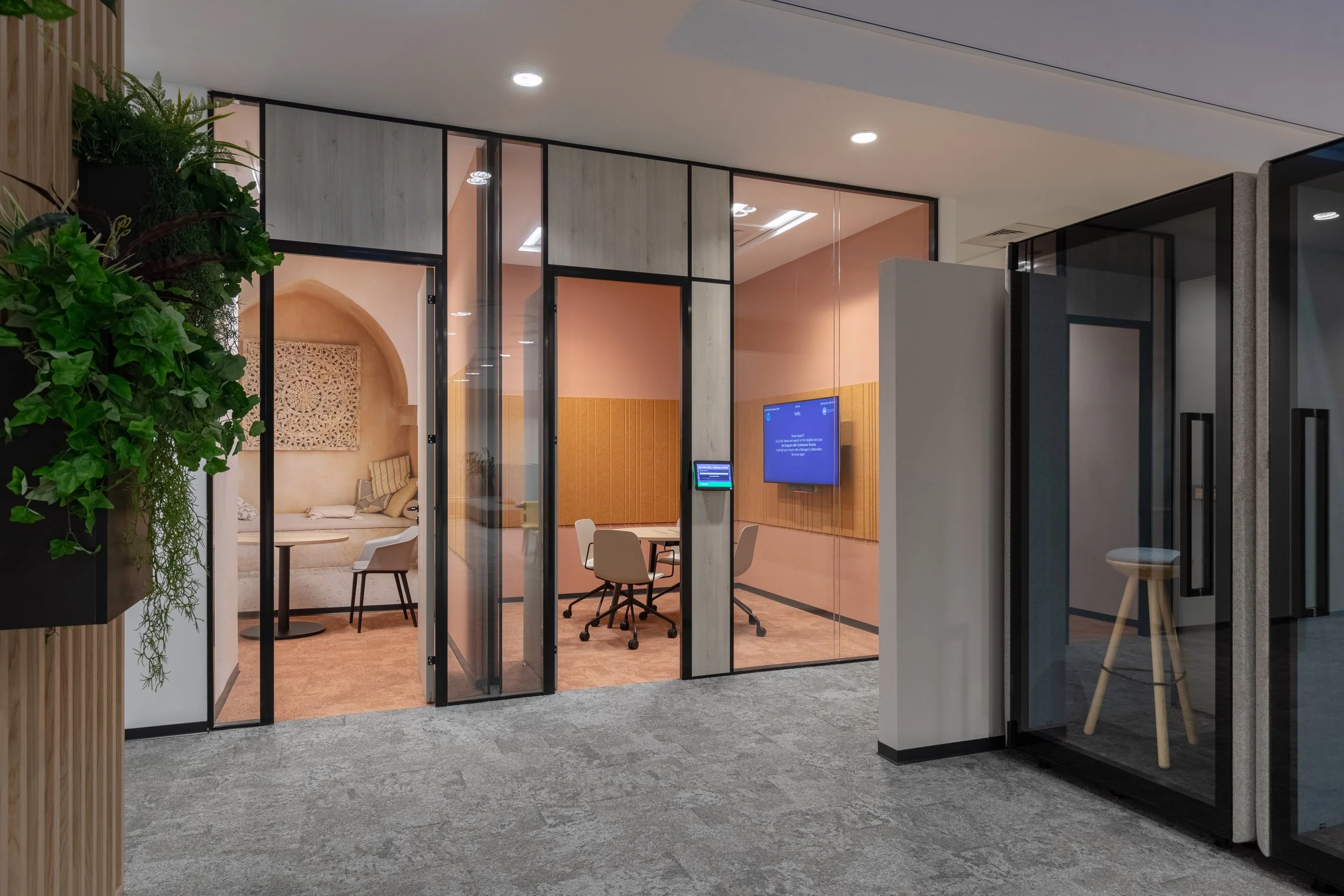 Modern office conference room with glass walls, a wall-mounted TV screen displaying a blue screen, and a small round table with chairs inside. A large plant with green leaves is nearby, and there's a small windowed private area with a decorative wall hanging and a table with chairs.
