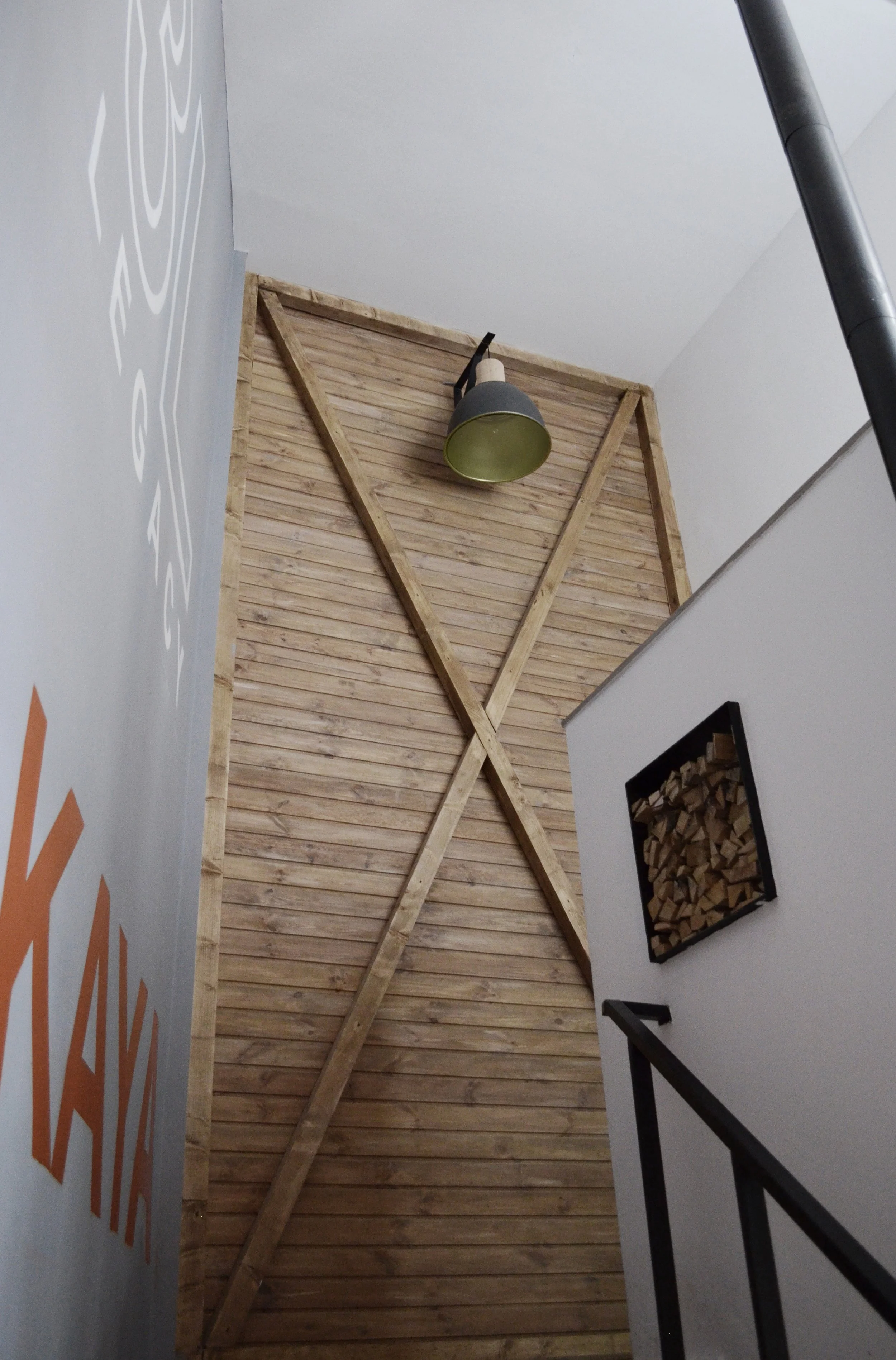 Looking up a staircase with a wooden wall, a black and green wall lamp, and a small square wood storage area on the white wall.