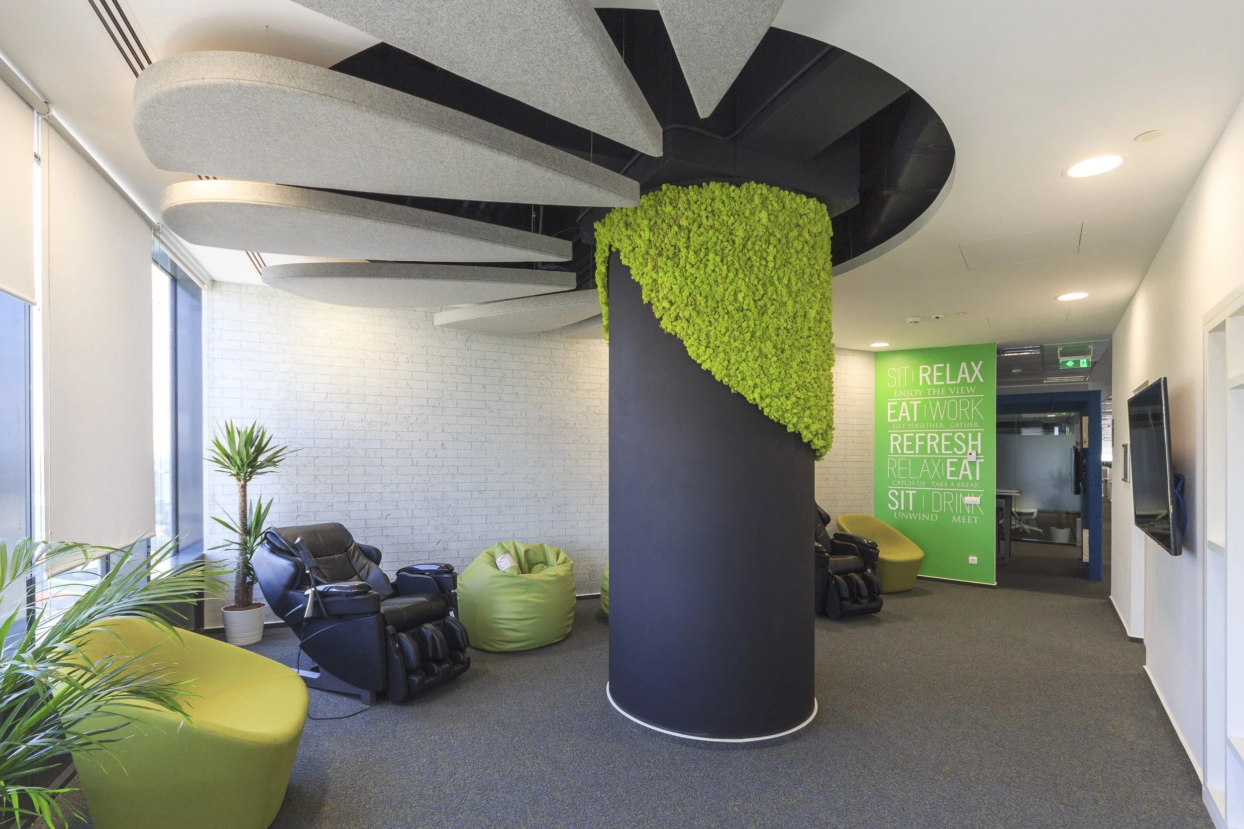 Modern lounge area with massage chairs, green bean bags, and plants, featuring a spiral staircase and a bright green wall with the words 'Sit, Relax, Eat, Work, Refresh, Relax, Eat, Sit, Drink' in white text.