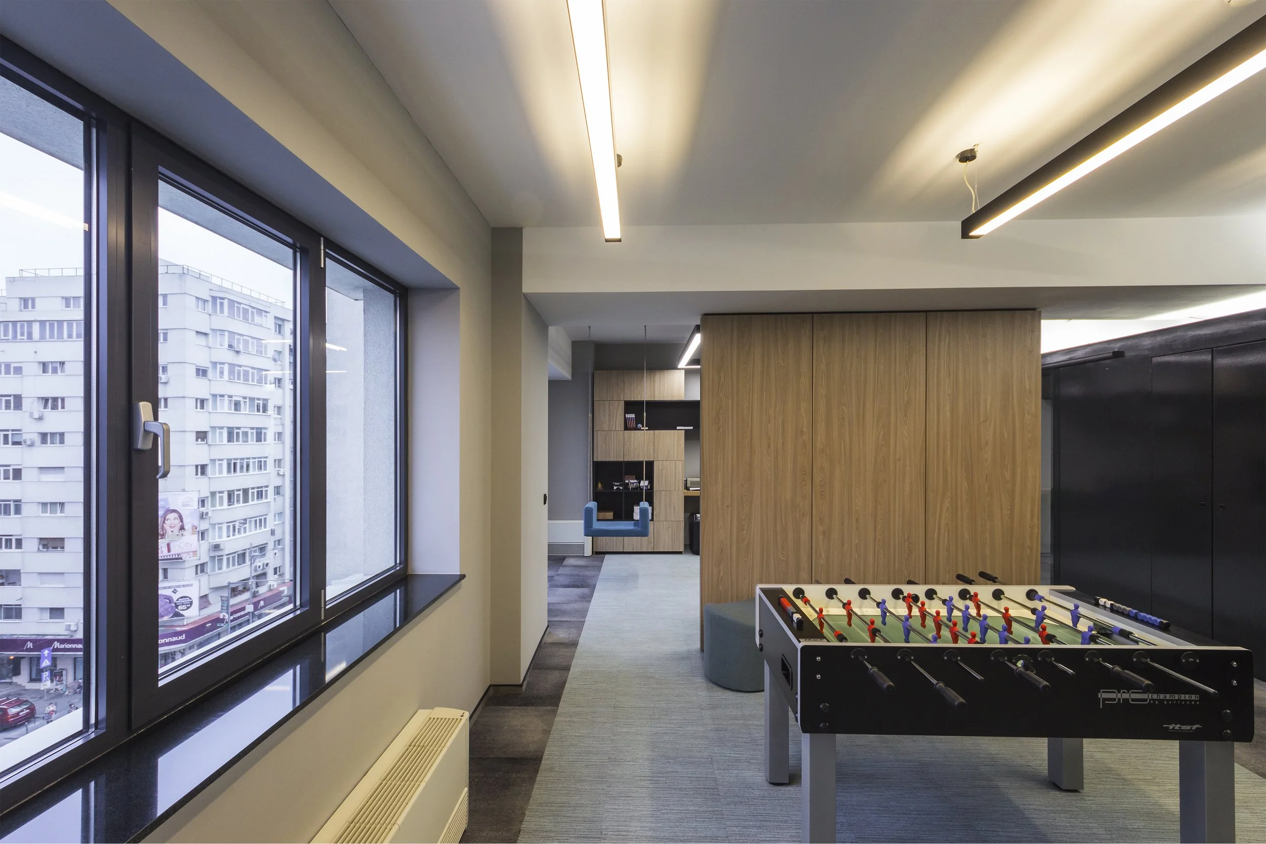 Modern office lounge with large windows showing city buildings, foosball table, wooden partition, and seating area with hanging blue chairs.