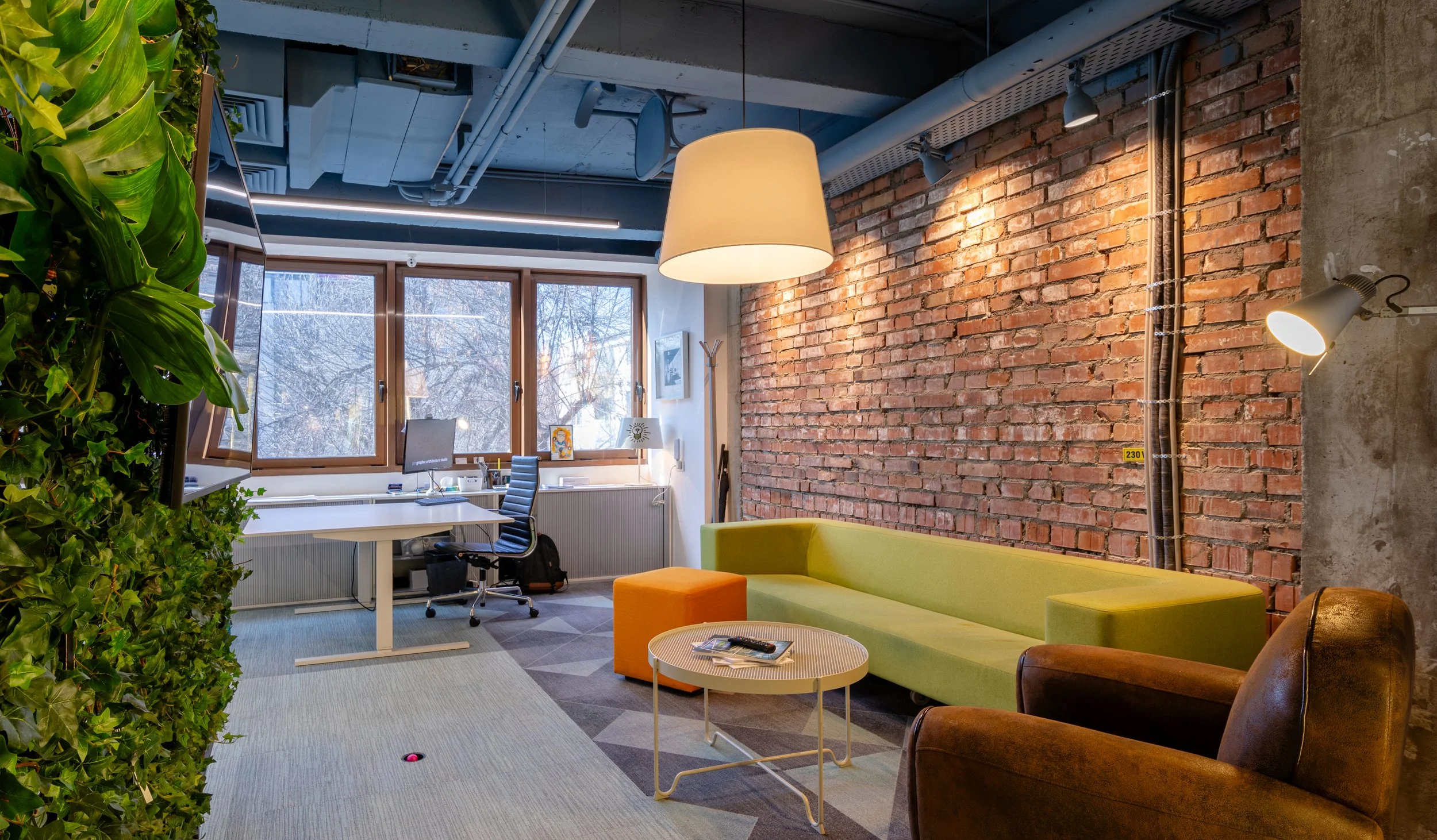 Modern office lounge with a green sofa, brown leather armchair, small round coffee table, and an orange ottoman, next to an exposed brick wall and large windows with winter trees outside.