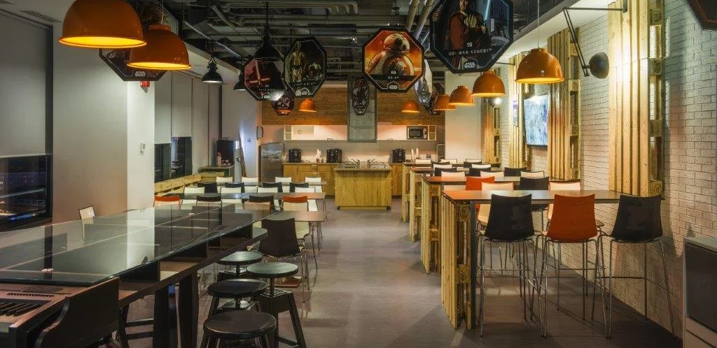 Empty modern cafe with high tables, bar stools, hanging orange pendant lights, wooden dividers, and a kitchenette area at the back.
