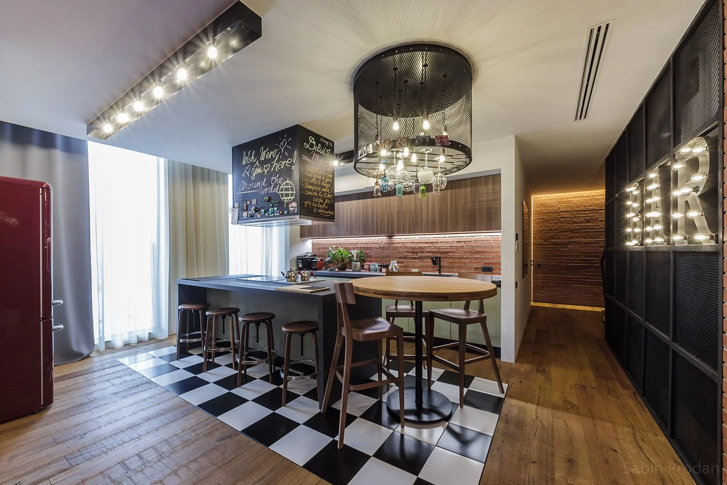 Modern kitchen and dining area with checkered black and white floor tiles, a circular wooden table, bar stools, brick wall, black metal and wood accents, and a chandelier with hanging glasses and exposed bulbs. Large window with curtains, and decorative lighting spelling "YORK" on a wall.