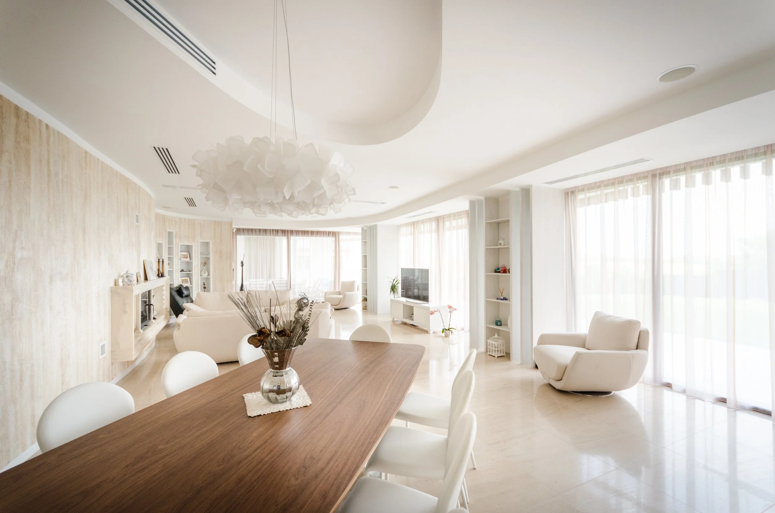 Bright, spacious living room with white furniture, including a sofa and armchair, large windows with sheer curtains, and a television on a white stand. A wooden dining table with white chairs and a decorative vase in the foreground.