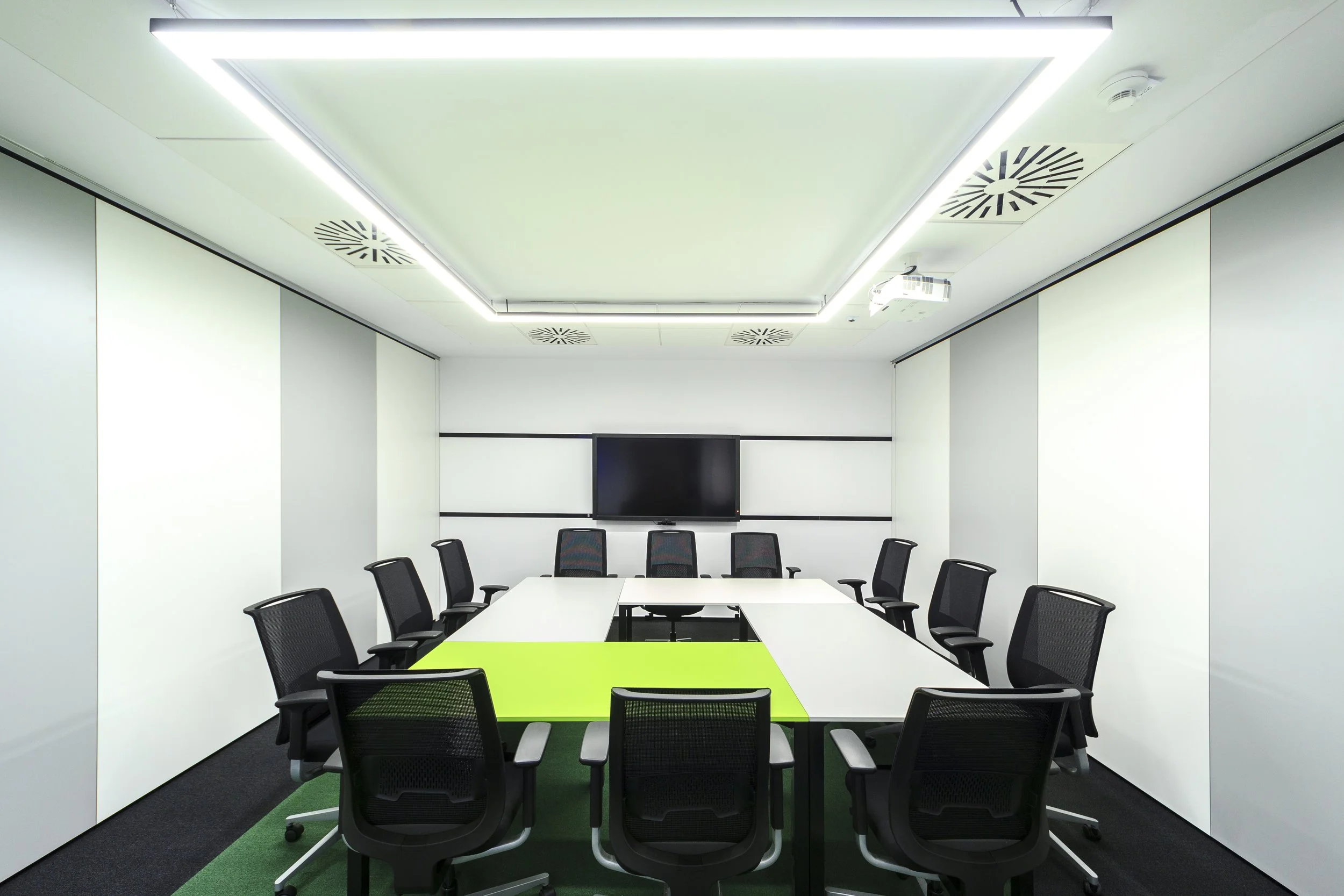 An empty modern conference room with white walls, a long white table with a green section, and twelve black office chairs around the table. A large flat-screen TV is mounted on the wall at the end of the room, and a ceiling with integrated lighting and air vents.