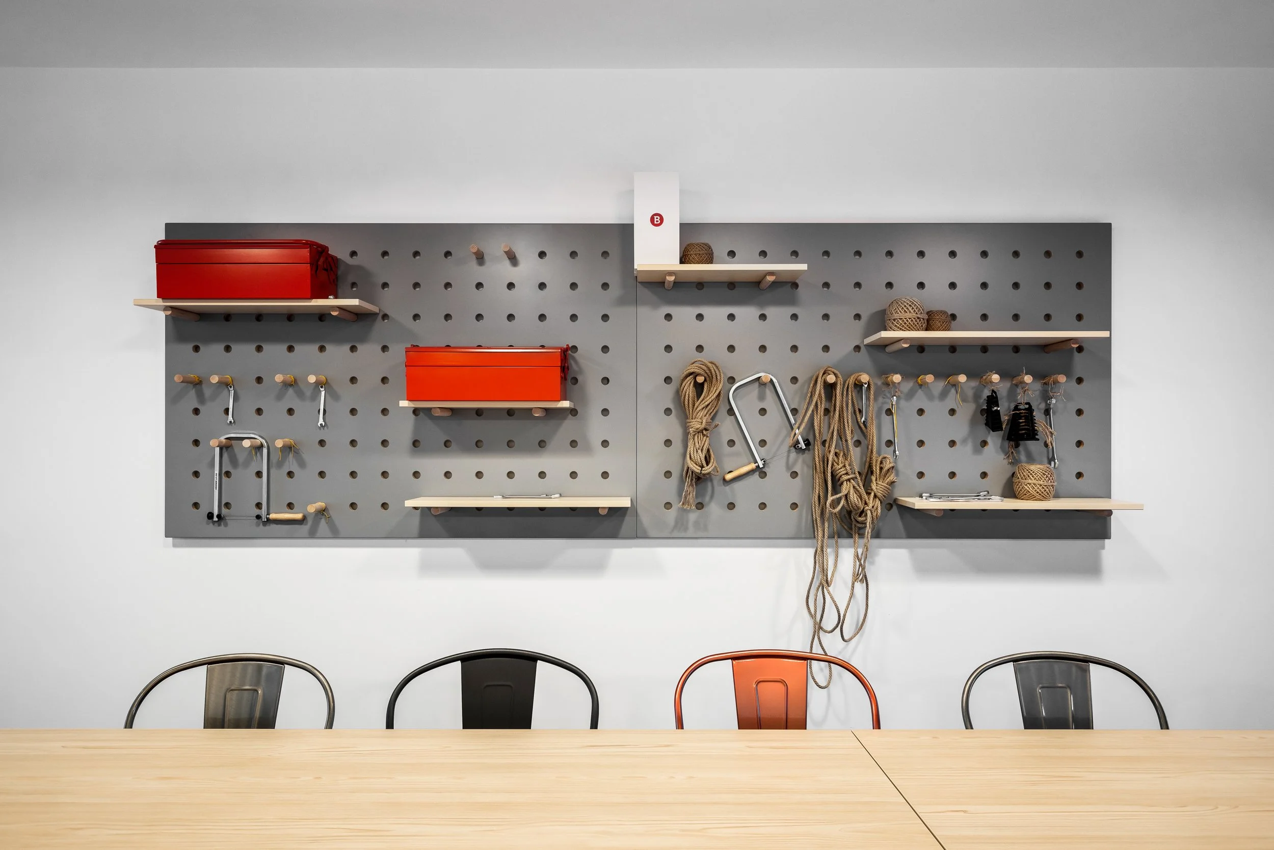 A workspace with a gray pegboard on the wall holding red boxes, ropes, hooks, and saws, with a wooden table and three metal chairs in front.