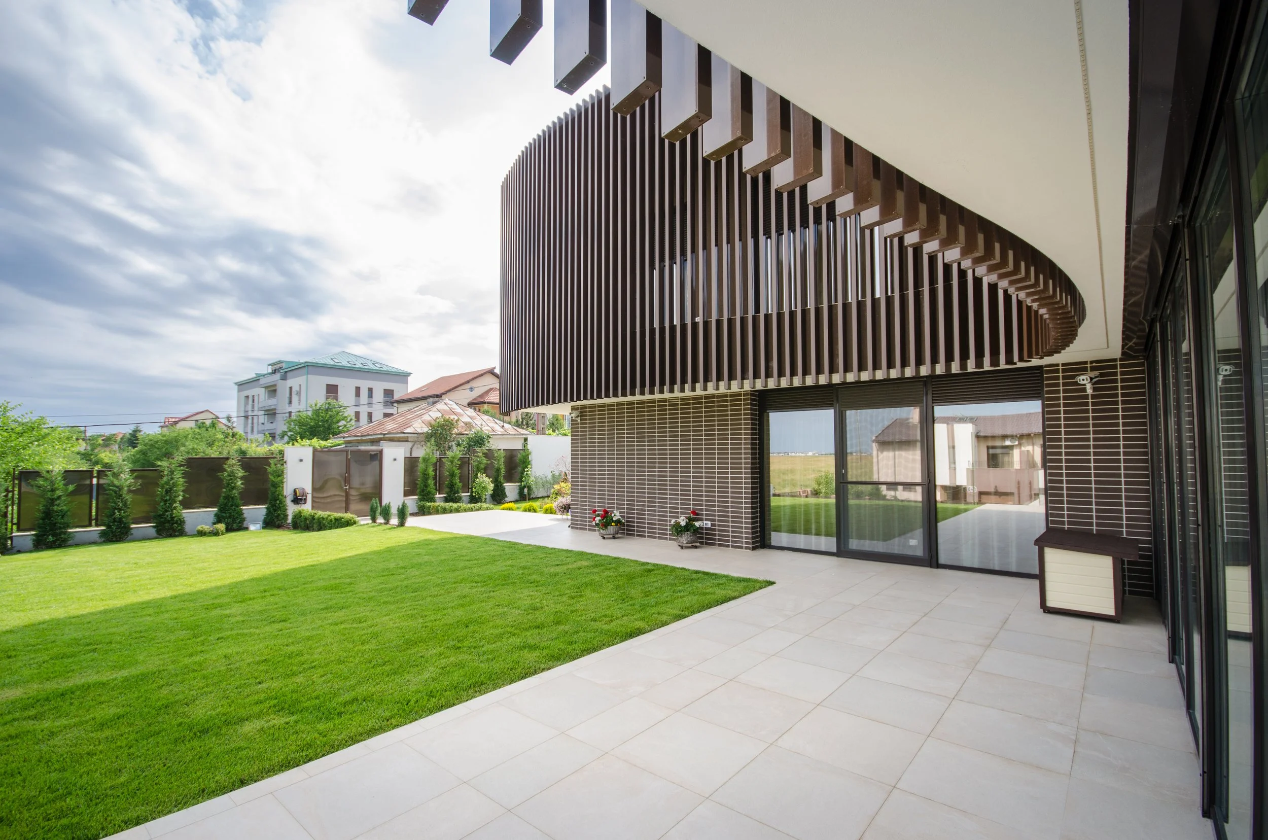 Modern building with a rounded, vertical wooden slat facade, large glass sliding doors, a spacious tiled patio, and a lush green lawn with small trees and shrubs.