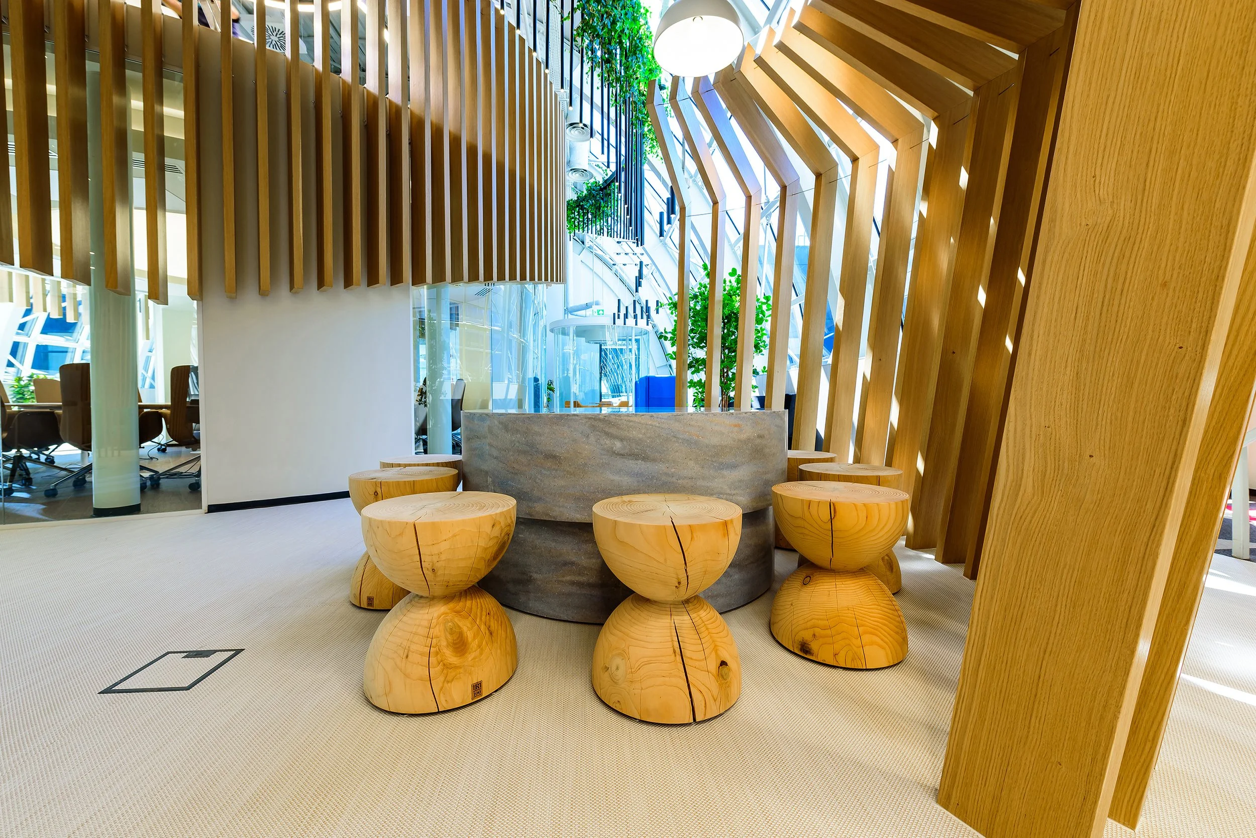 Modern interior space with wooden furniture, including tables and a curved bench, accented with wooden slat wall dividers and decorative greenery.