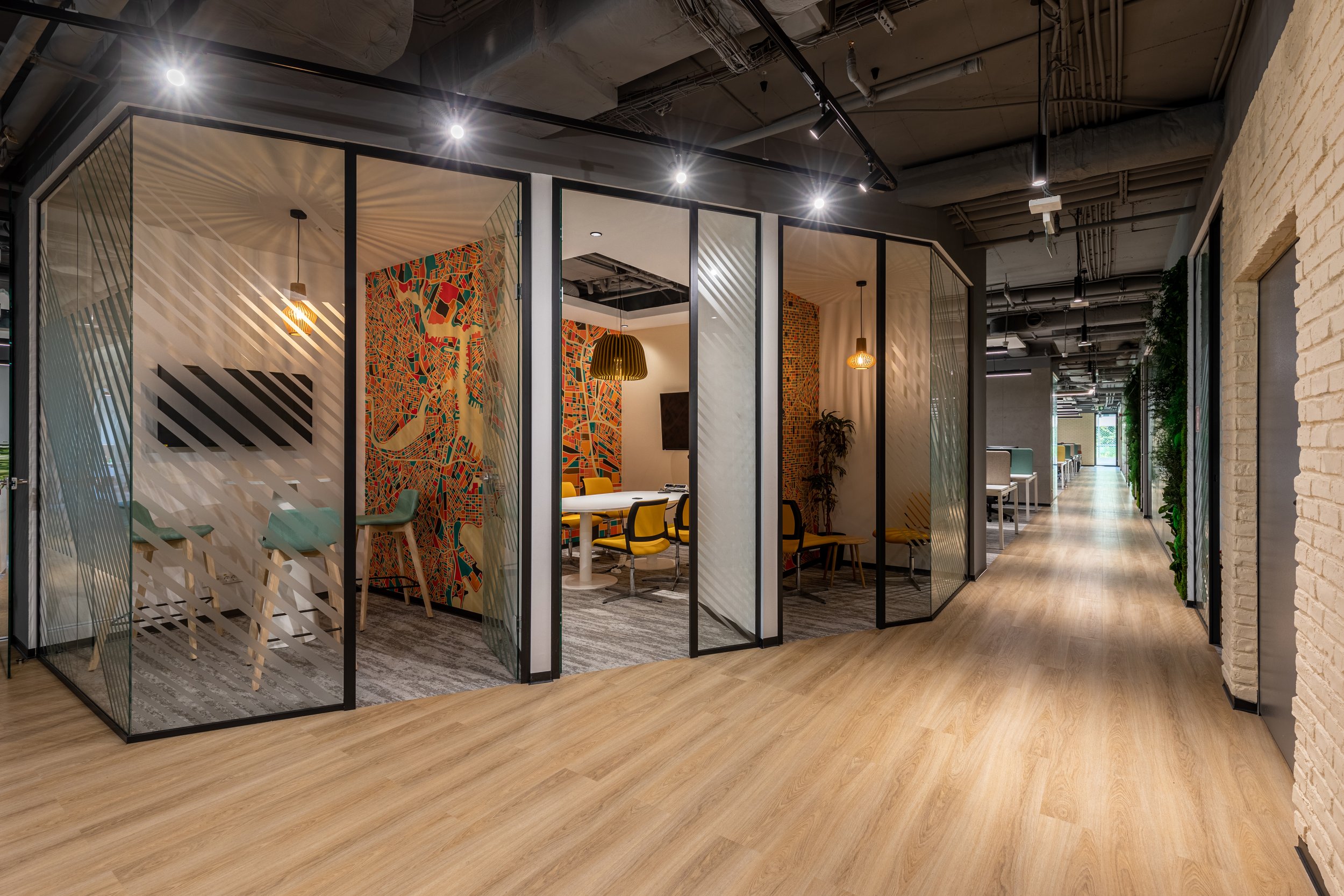 Modern office corridor with private glass meeting rooms on left and open desks along the right wall, wooden flooring, exposed ceiling features, and a bright window at the end.