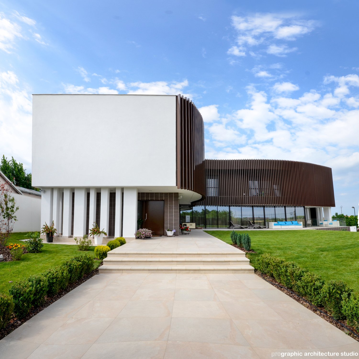 Modern house with a white and brown curved facade, front garden with shrubs and flowers, and a patio with lounge chairs under a partly cloudy sky.