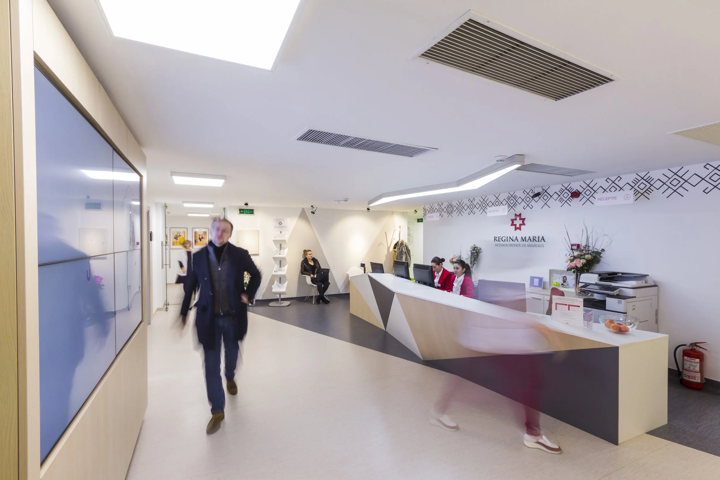 Reception area of Regina Maria healthcare facility with two receptionists at desks, a man walking, and two seated women, modern decor with white walls, floral arrangements, and digital displays.