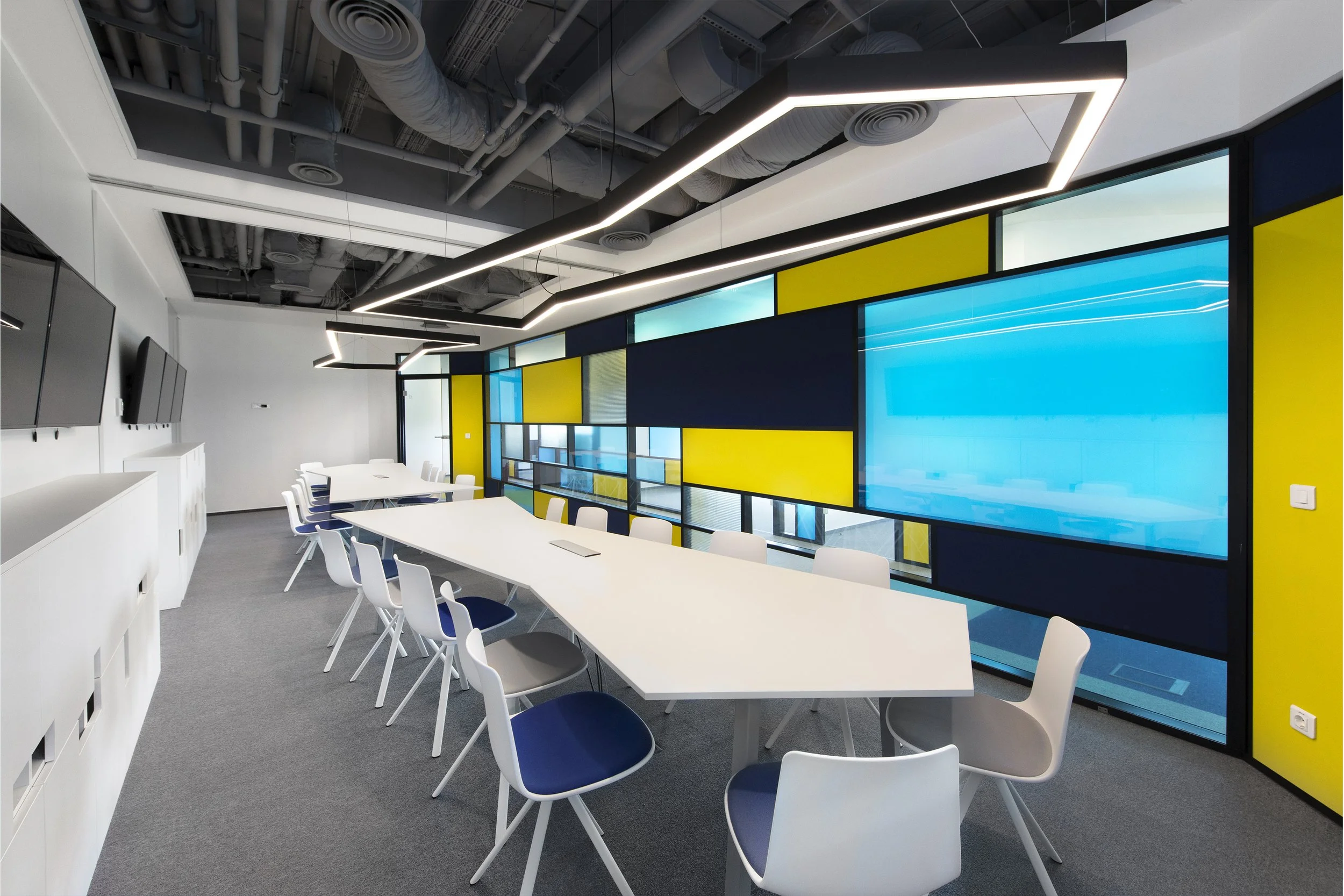 Modern conference room with a large white table, white chairs with blue seats, yellow and blue accent wall, and ceiling with exposed ductwork and linear LED lighting.