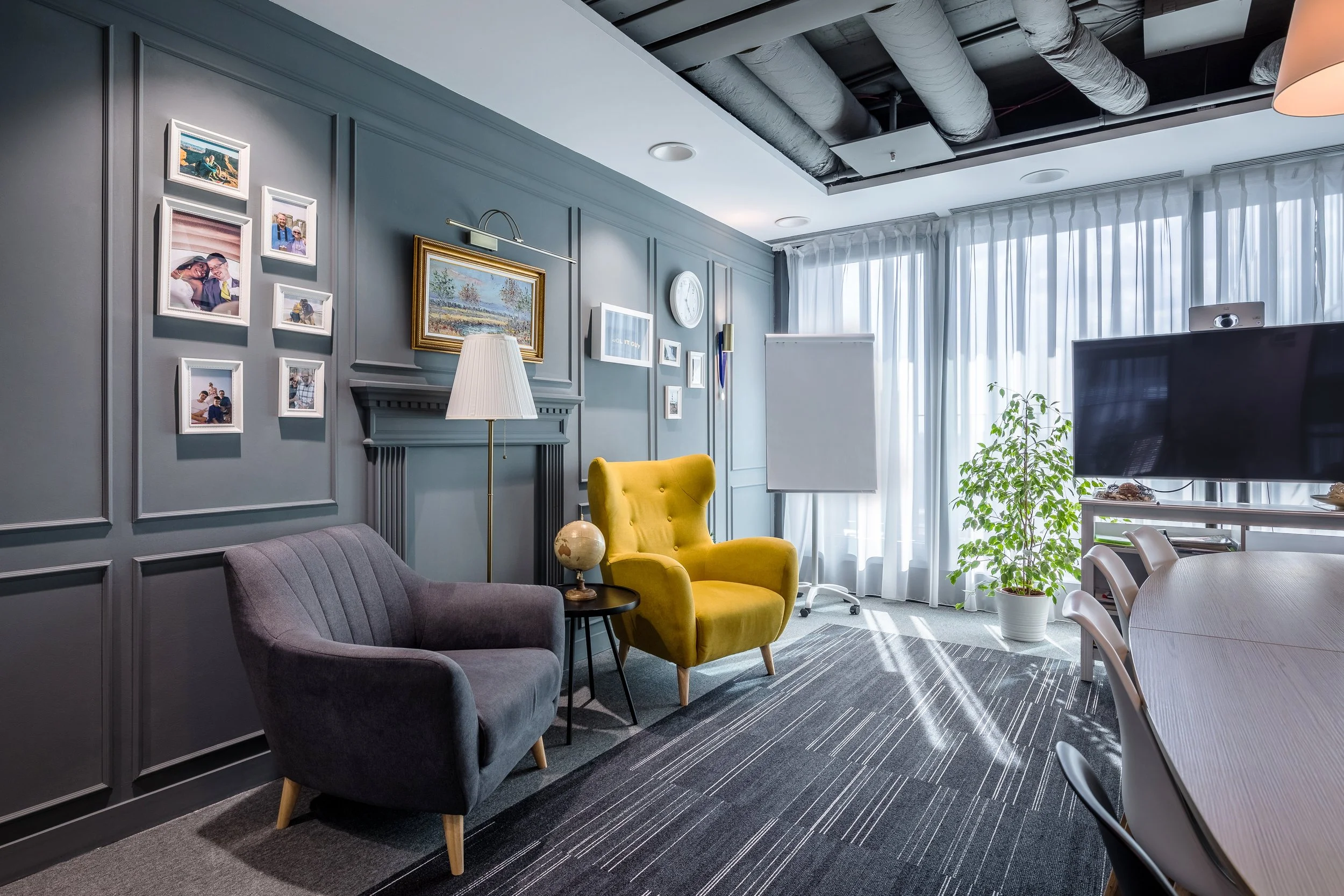 Interior of a modern living room with gray wall paneling, a yellow armchair, a gray loveseat, a floor lamp, wall art, a large window with white curtains, a potted plant, a whiteboard, and a TV.