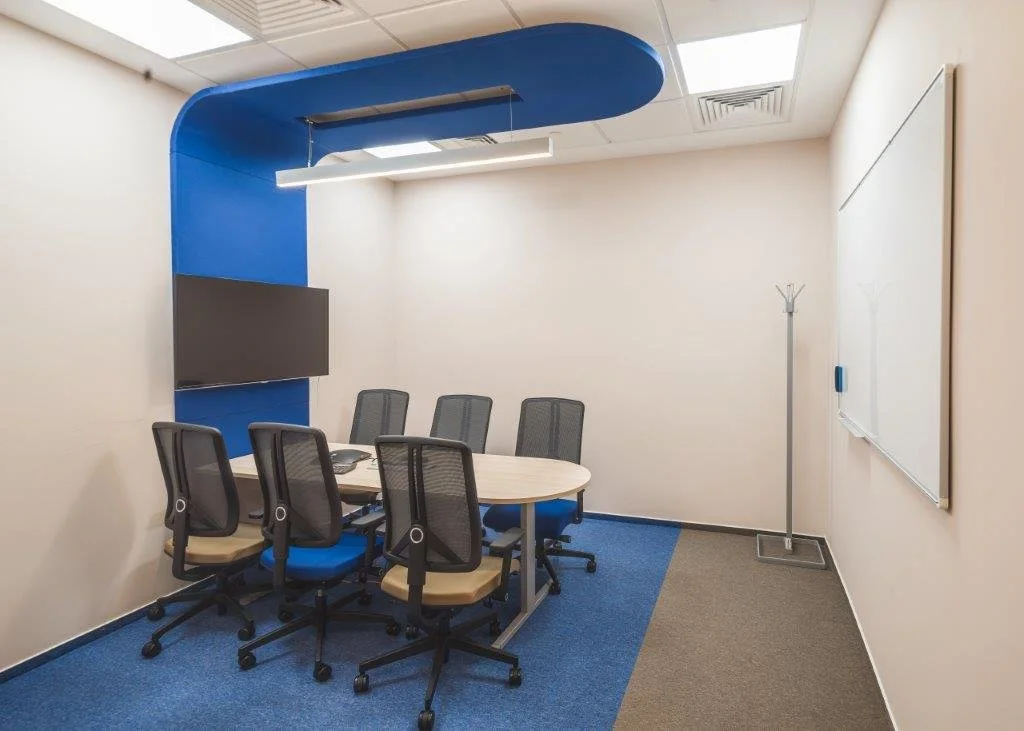 Small conference room with a round table, six black office chairs, a whiteboard on the right wall, a coat rack on the right corner, a flat-screen TV on the blue wall behind the table, and ceiling lights.
