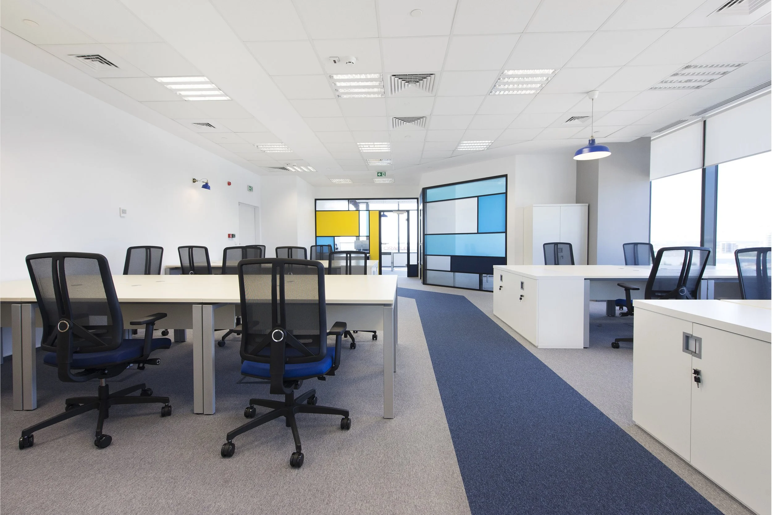 Empty modern office space with white desks, black office chairs, and a colorful blue and yellow wall divider, illuminated by natural light through large windows.