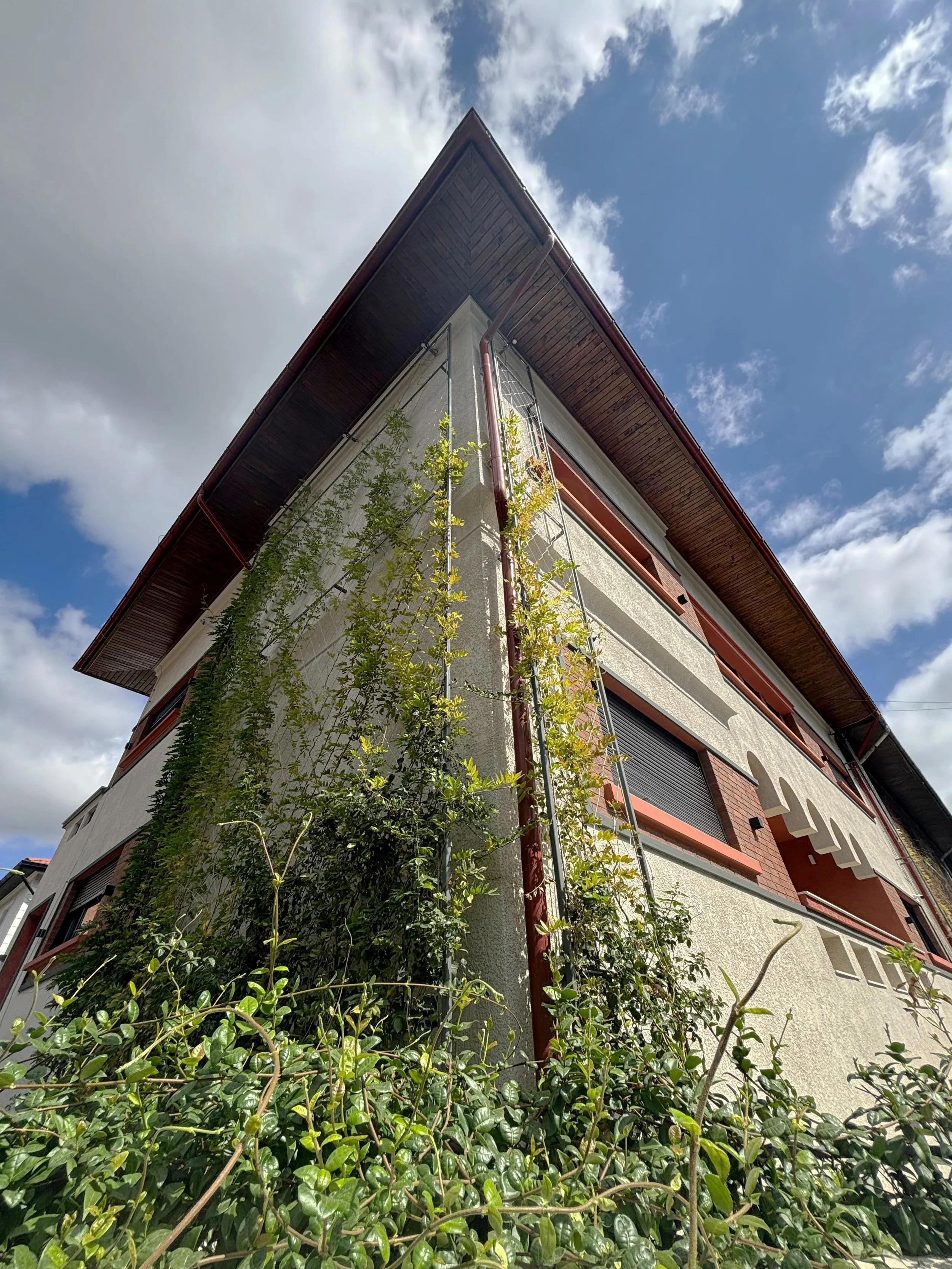 Low-angle view of a tall residential building with a sloped roof, surrounded by greenery and a partly cloudy sky.