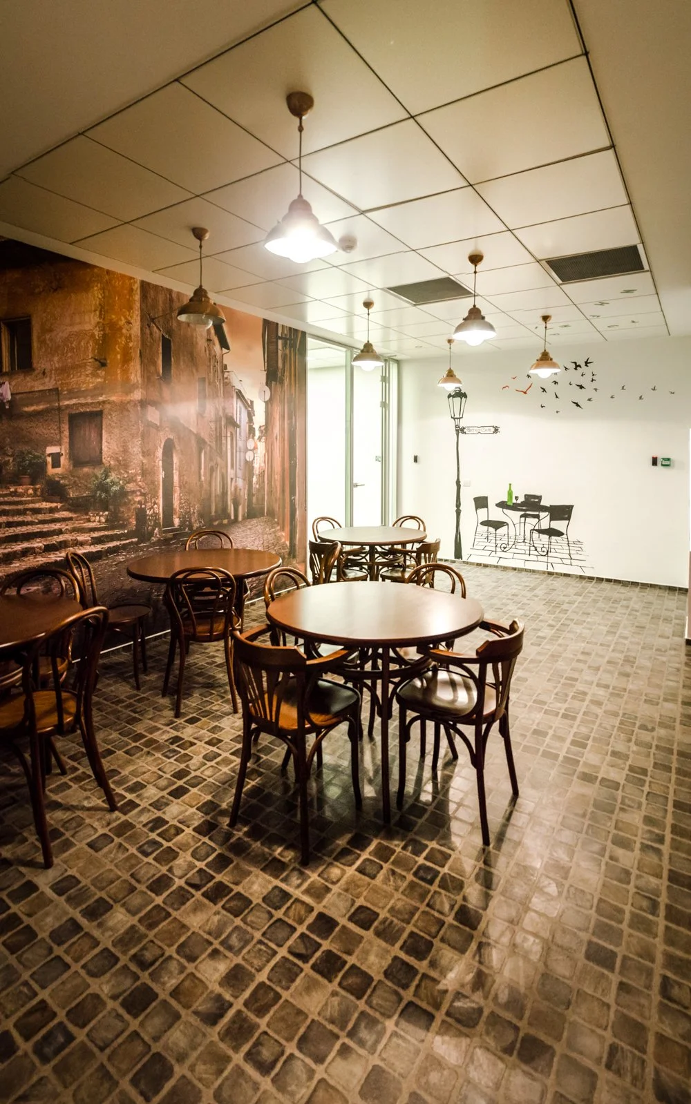 Interior of a restaurant with round tables and chairs, a large mural of an old street scene on one wall, and a white wall with black line art of outdoor furniture, a street lamp, and birds.