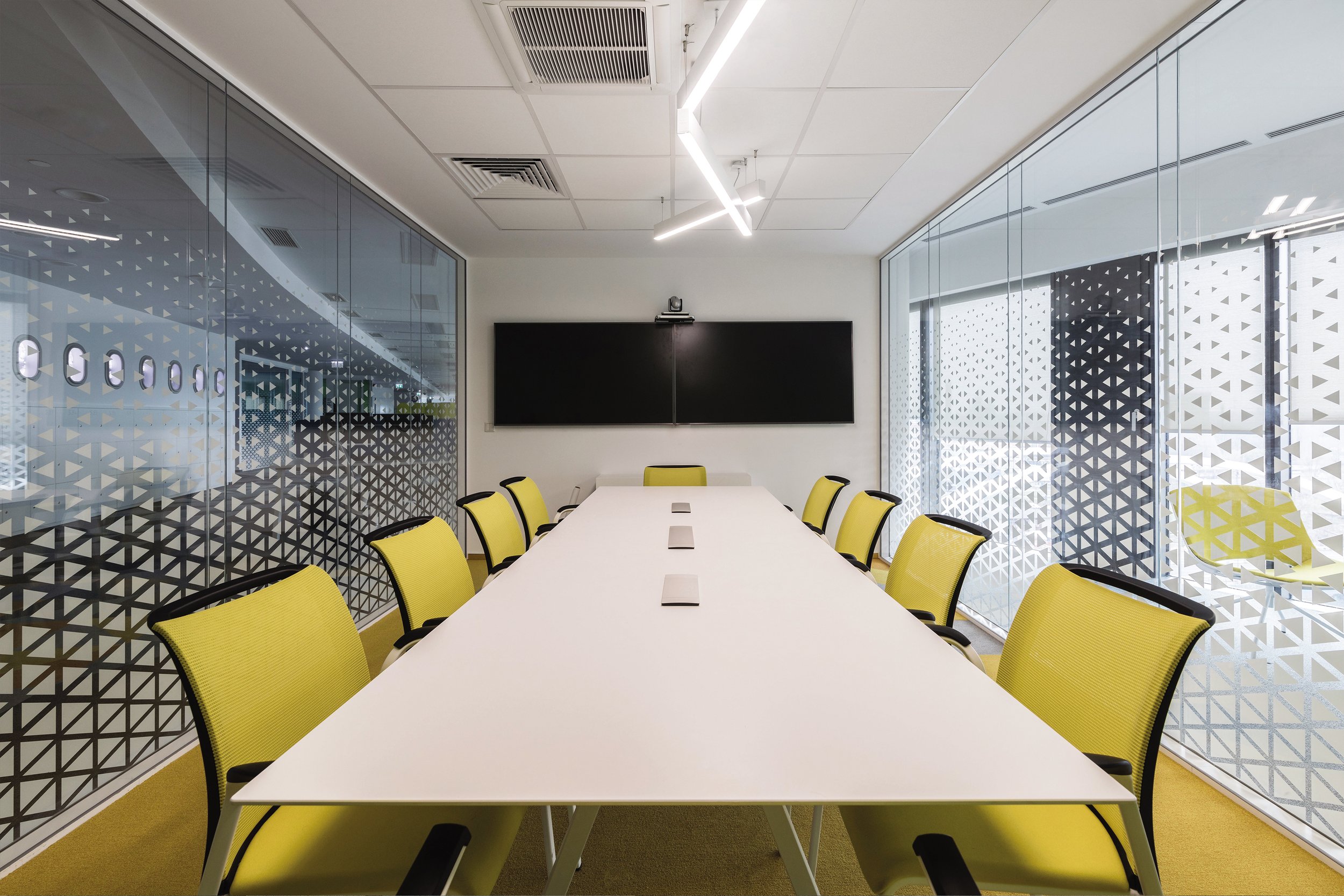 Modern conference room with a long white table, yellow chairs, a black screen on the wall, glass walls with geometric patterns, and ceiling lights.