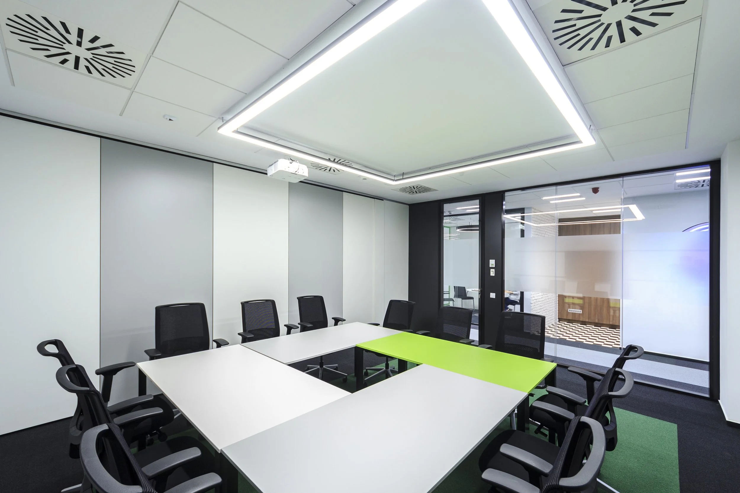 Modern conference room with U-shaped white tables, black office chairs, glass wall, and ceiling lights.
