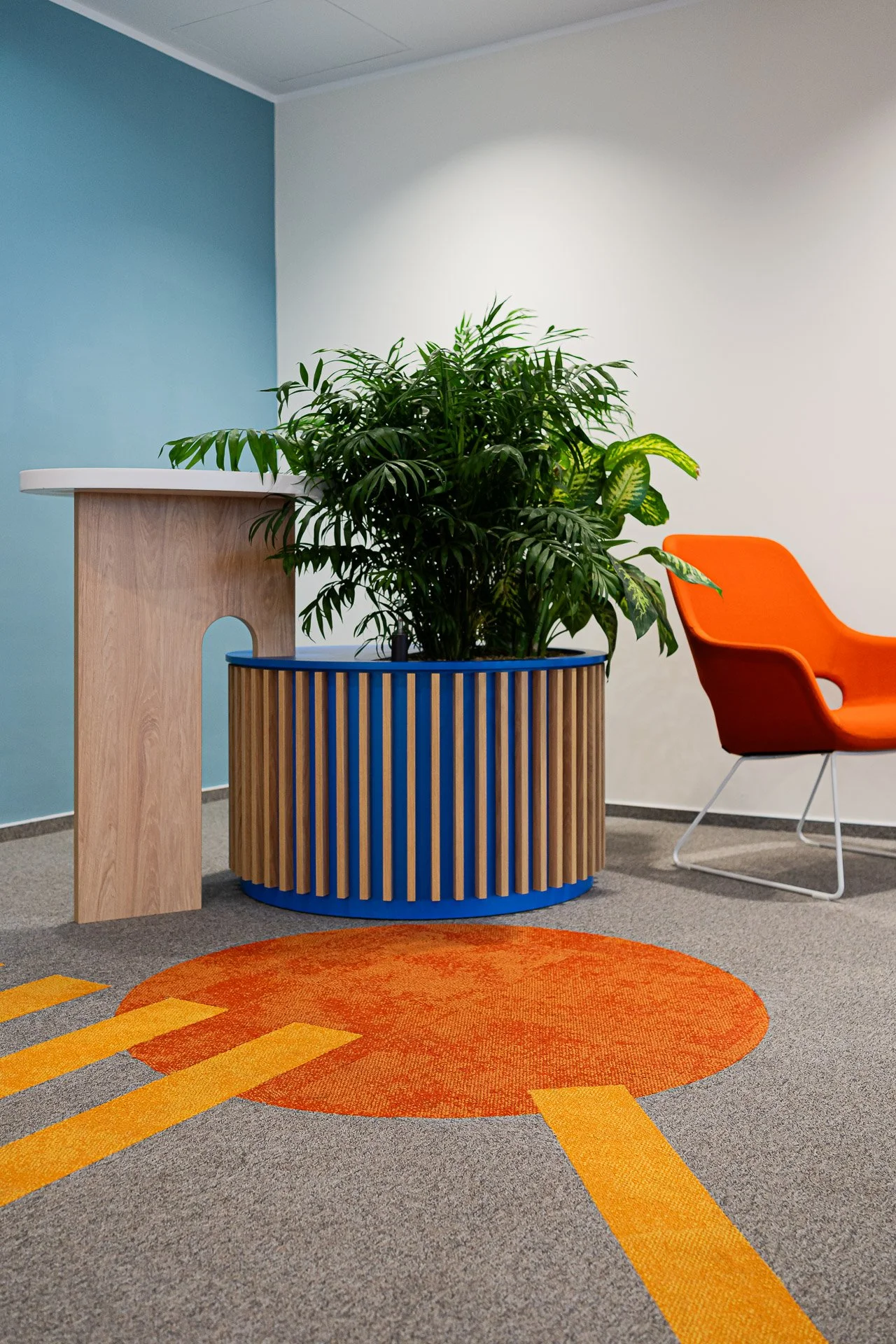 An indoor office corner with a round blue wooden planter filled with a large green leafy plant, a modern orange chair, a wooden reception counter, and a colorful carpet with orange and yellow geometric patterns.