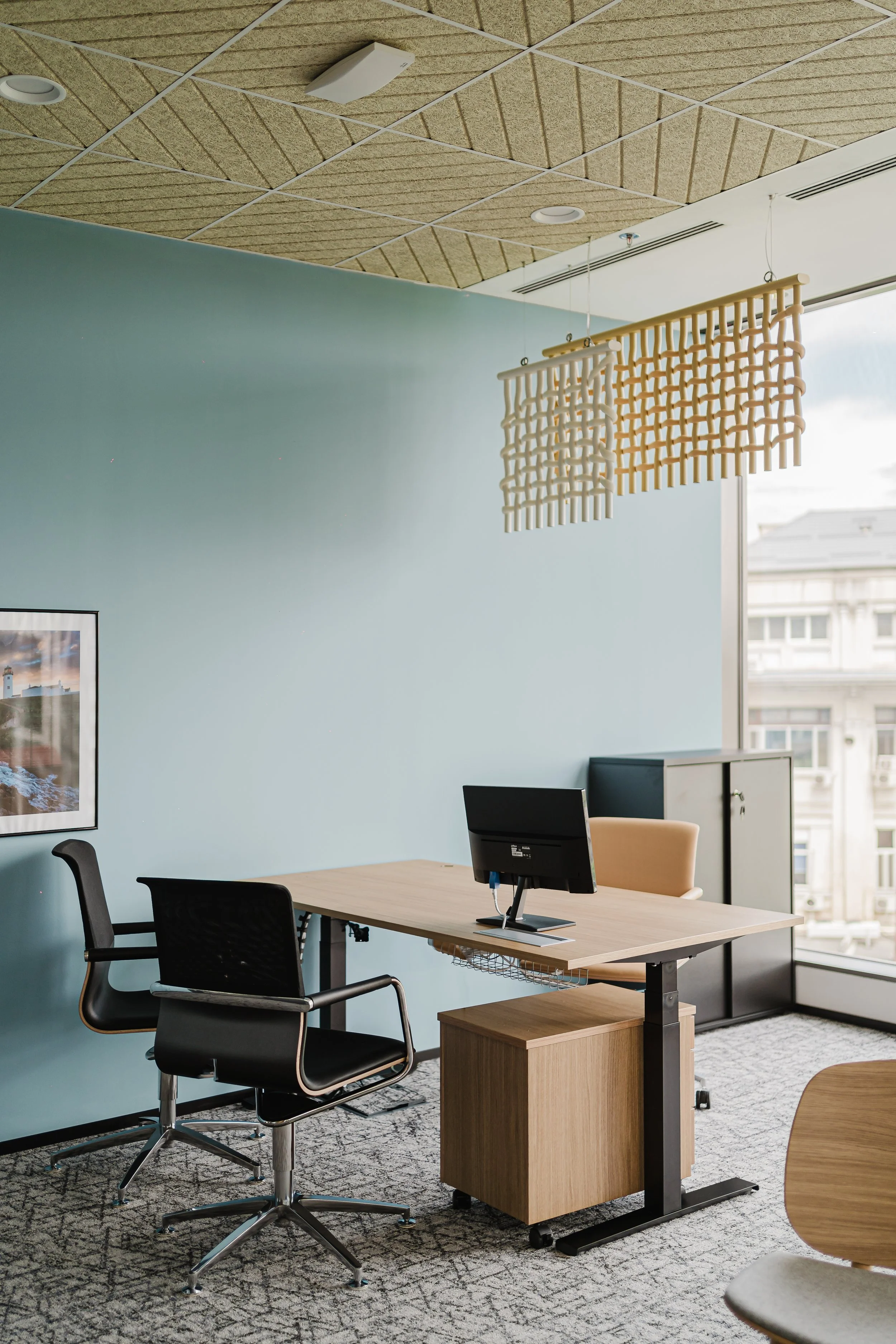 An office with a desk, two chairs, a computer, a small cabinet, a large window, framed artwork, and a modern hanging light fixture.
